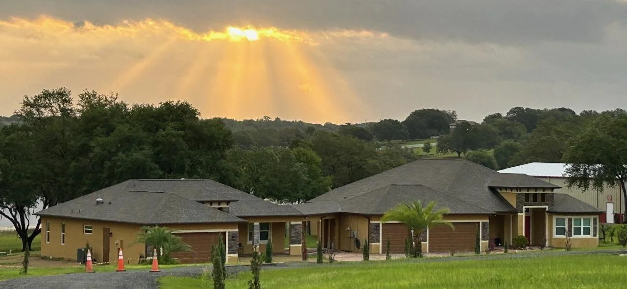 Large modern house with a lawn, surrounded by trees, under a cloudy sky with sun rays breaking through the clouds.