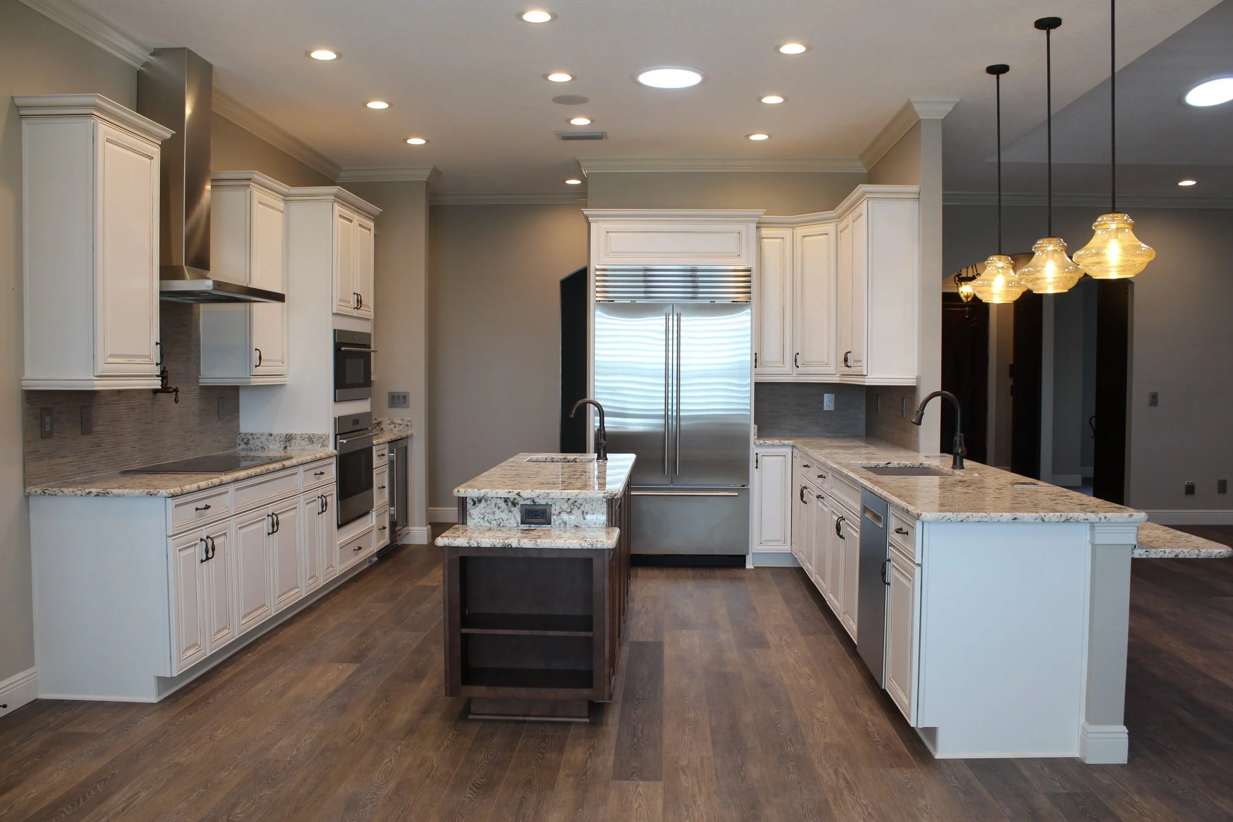 Modern kitchen with white cabinets, granite countertops, stainless steel refrigerator, island with sink, wooden flooring, and pendant lighting.