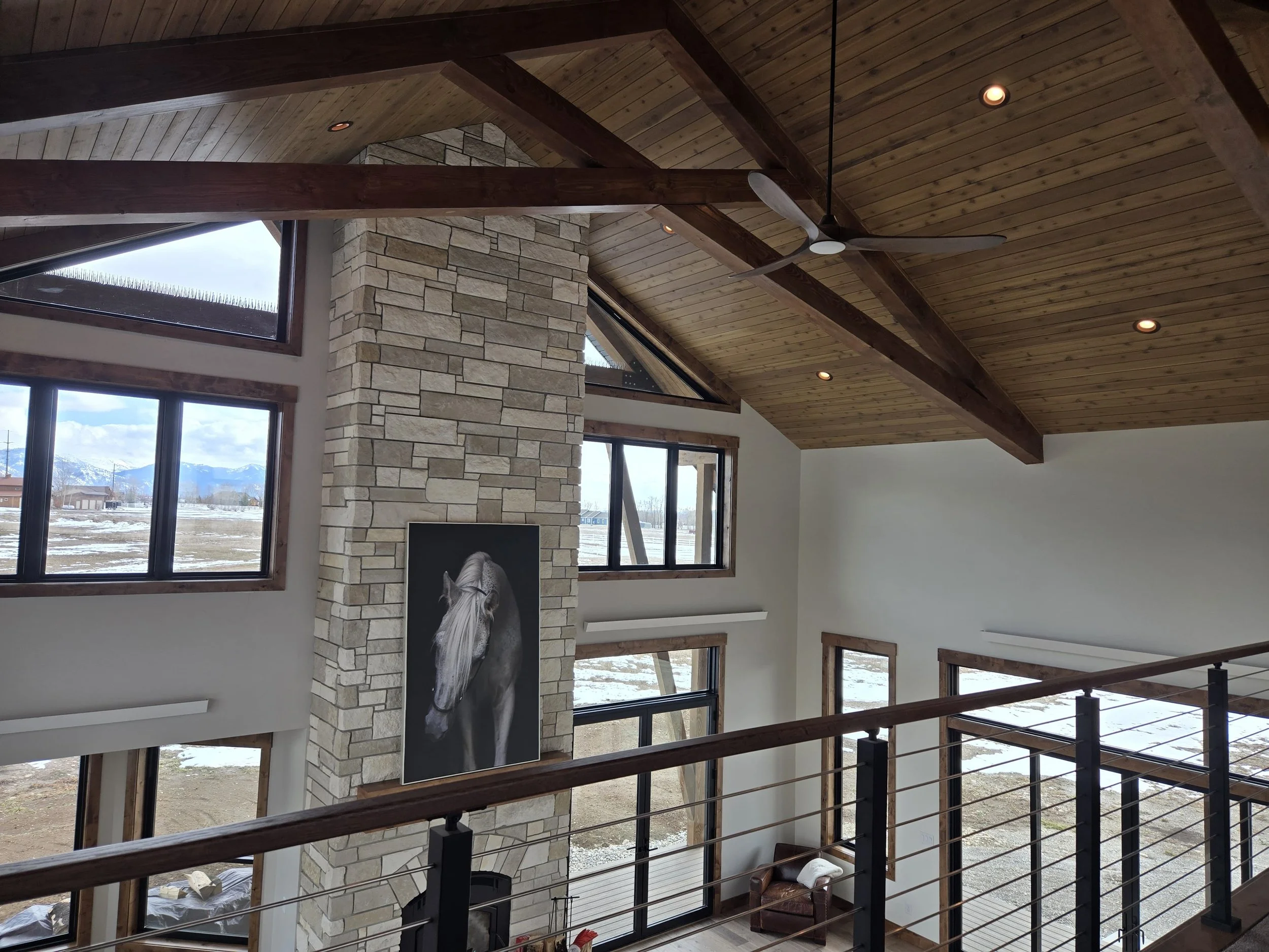 Interior of a modern house with a high vaulted wooden ceiling, large windows, a stone fireplace with a horse portrait, and a balcony railing.