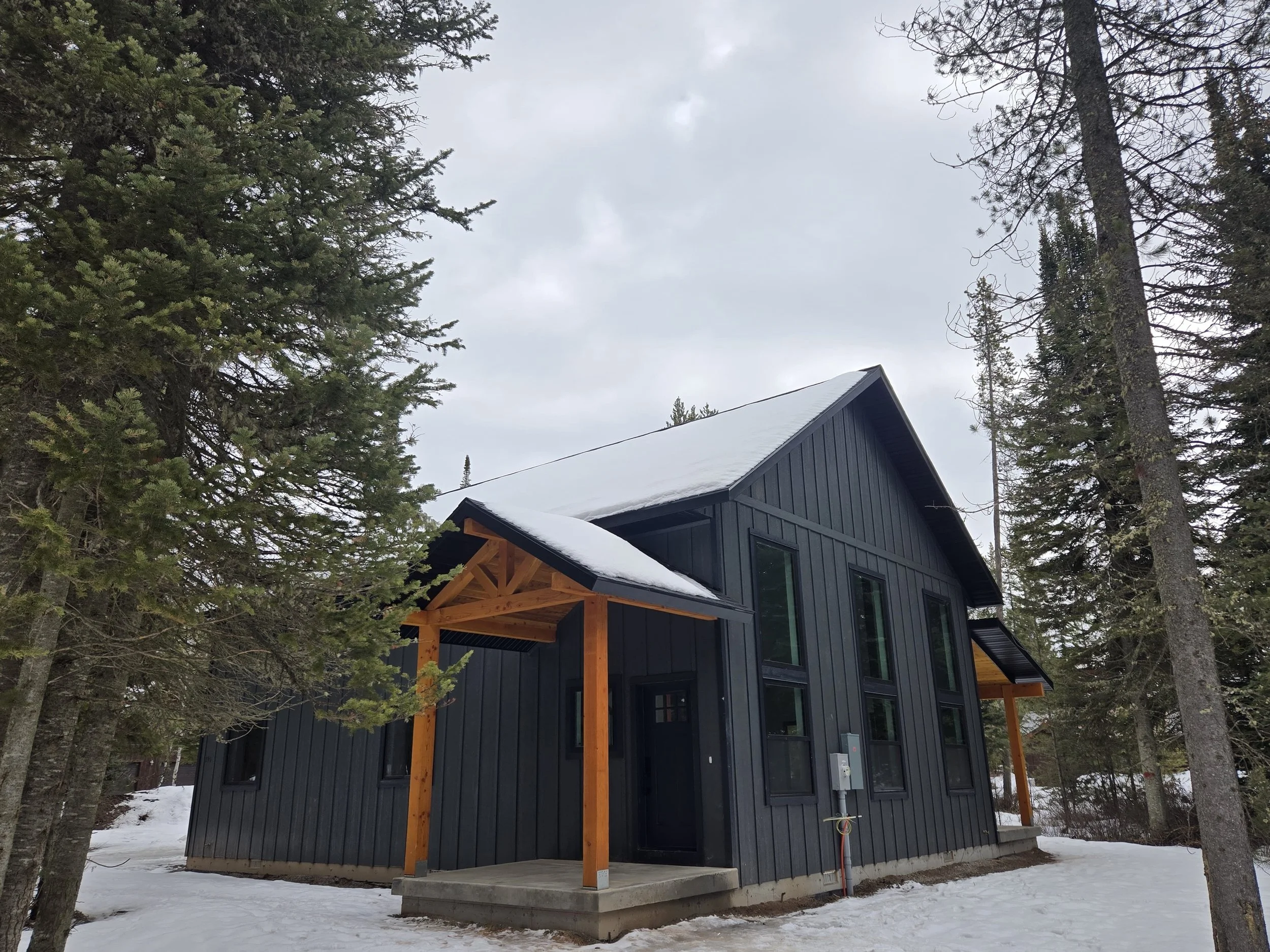 A modern black house with large windows and wooden porch supports, situated among snow-covered ground and tall evergreen trees on a cloudy day around a house in Alpine WY.