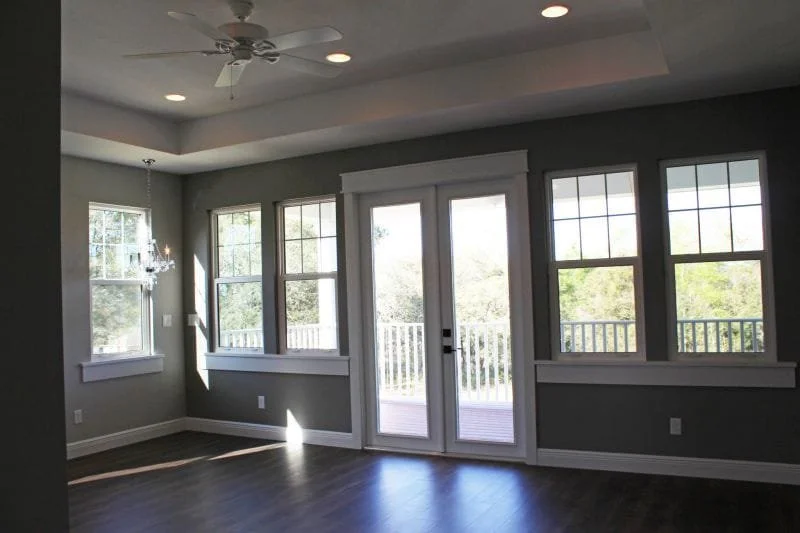 Empty living room with large windows and glass double doors opening to a balcony, gray walls, dark wood flooring, and recessed ceiling lights.