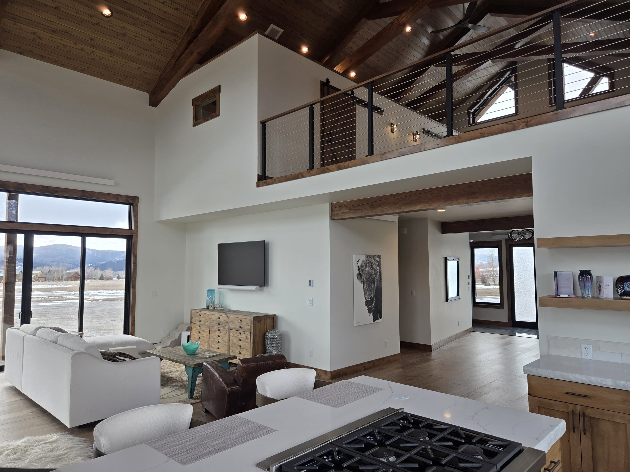 Interior view of a modern, open-concept living space with white walls, wooden accents, vaulted ceiling, and large windows showing a mountain landscape. Features include a white sofa, a wooden cabinet, a wall-mounted TV, and a kitchen counter with sto