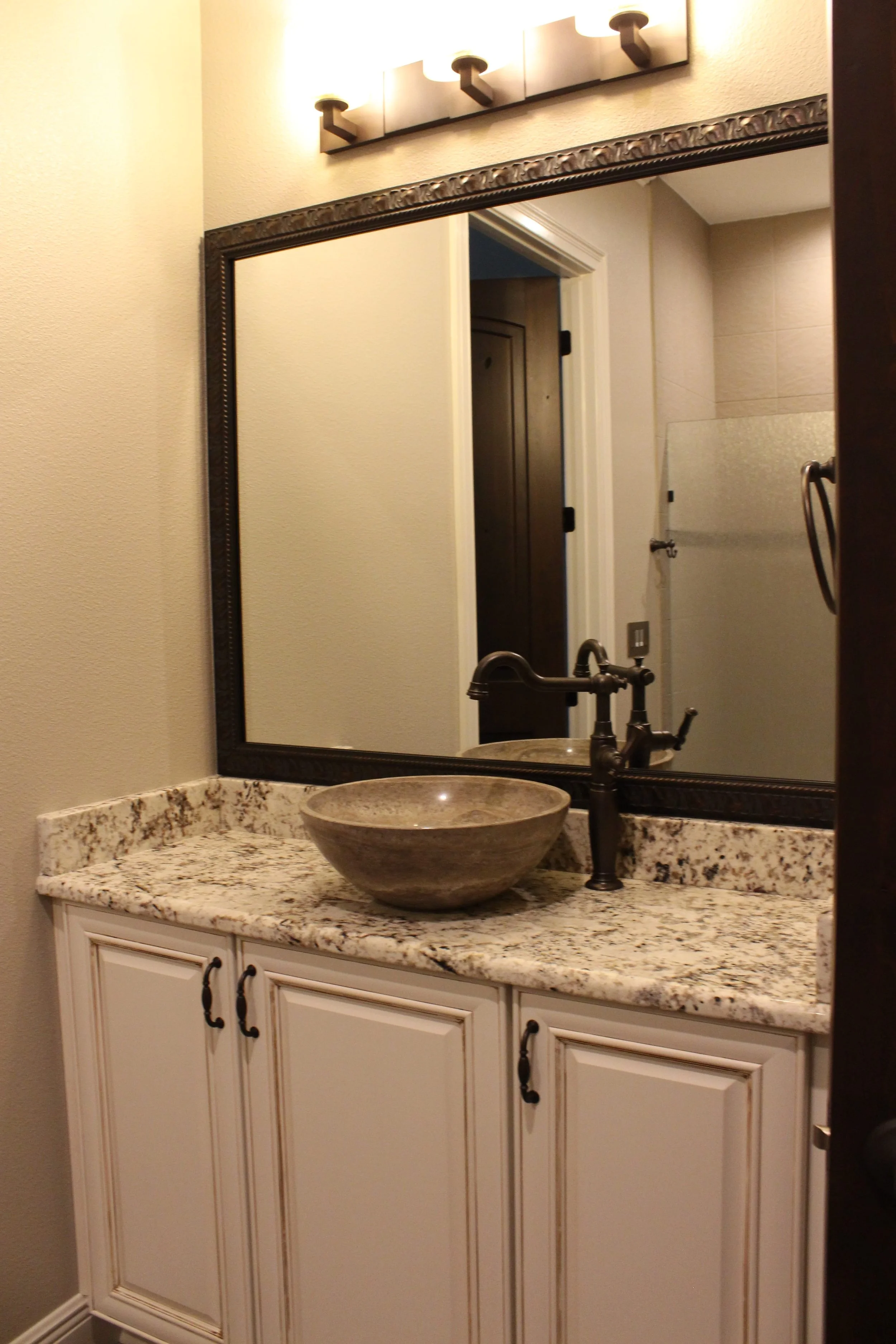 Bathroom vanity with a granite countertop, a stone vessel sink, a bronze faucet, a large framed mirror, and a light fixture above the mirror.