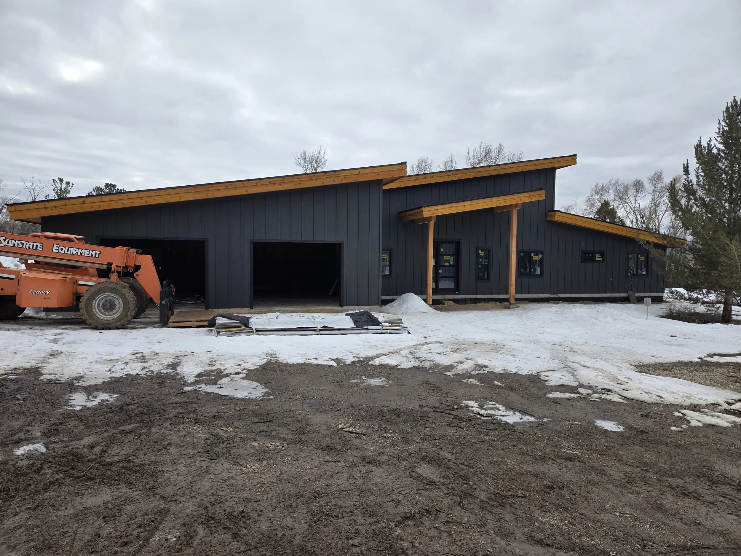 A modern black wooden house under construction with large garage doors and multiple small windows, surrounded by snow on the ground, with construction equipment nearby.