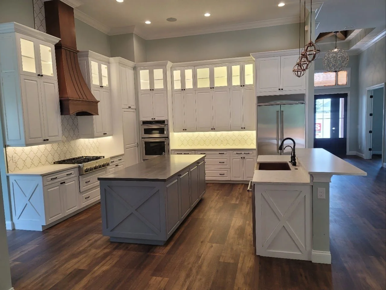 Modern kitchen with white cabinetry, a central island with a gray countertop, a built-in double oven, stainless steel refrigerator, and brown wooden flooring.