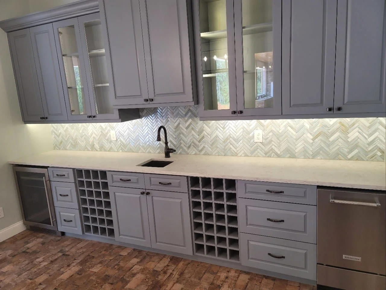 Kitchen with gray cabinets, marble countertop, herringbone tile backsplash, wine storage, small fridge, and a sink with a black faucet.