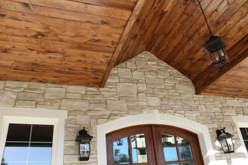 Close-up of a rustic house exterior showing a stone wall, wooden ceiling, and two wall-mounted lanterns around a wooden arched door with glass panes.