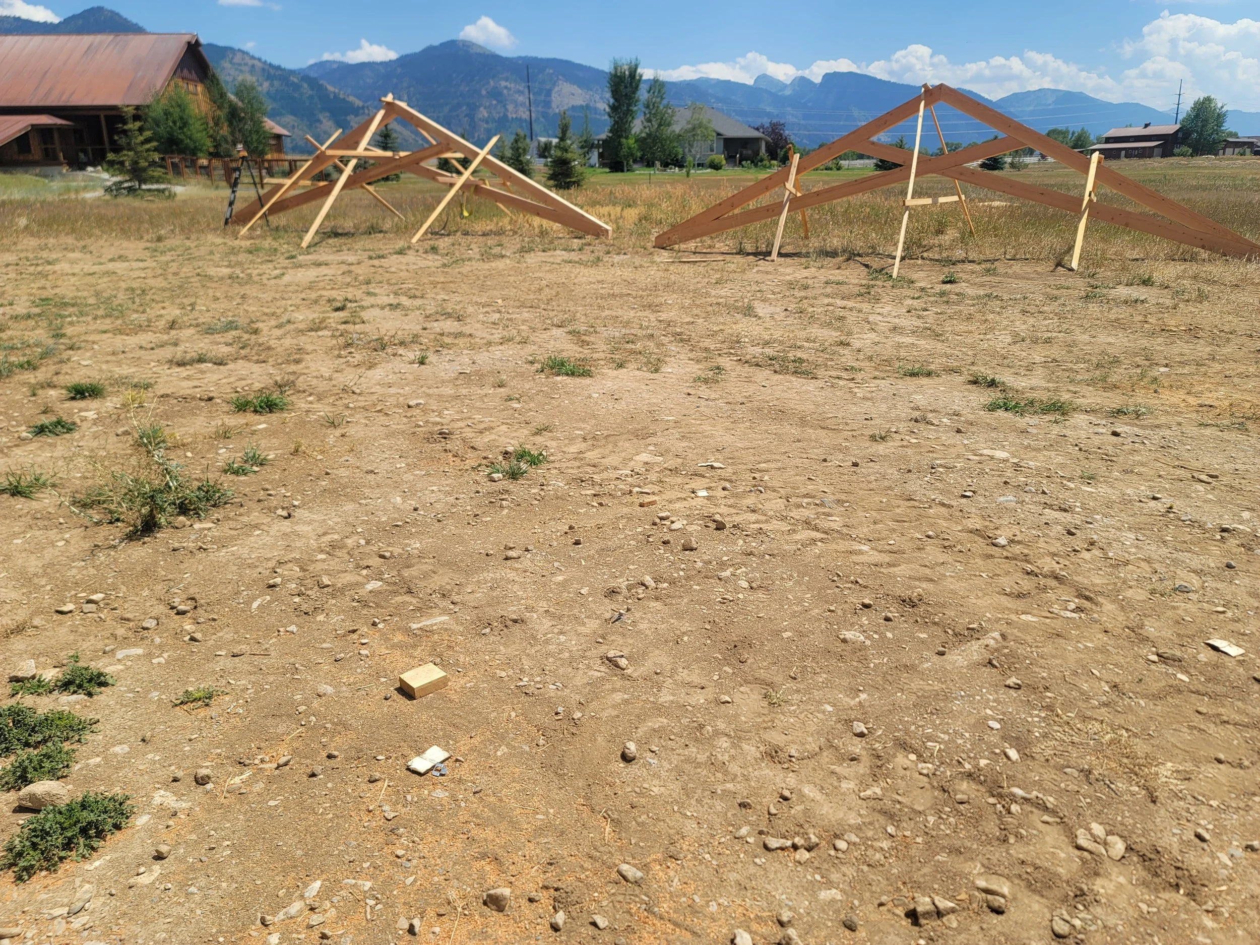Construction site with partially assembled wooden structures in a dirt field, with mountains and houses in the background.
