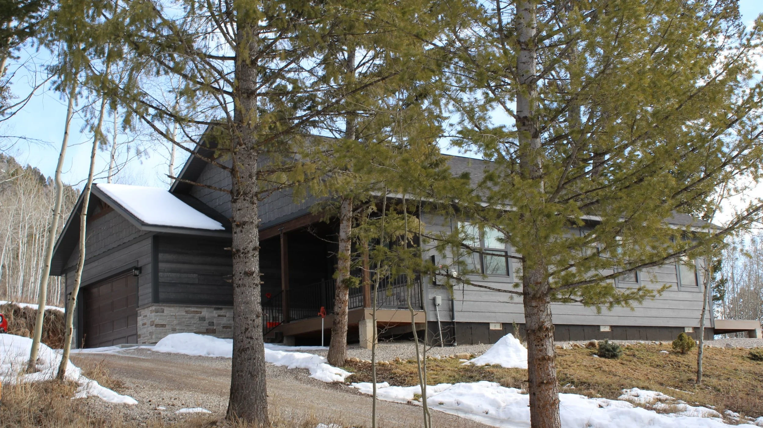 A modern house with gray siding, a stone accent on the lower wall, and a brown garage door is surrounded by trees and patches of snow on the ground.