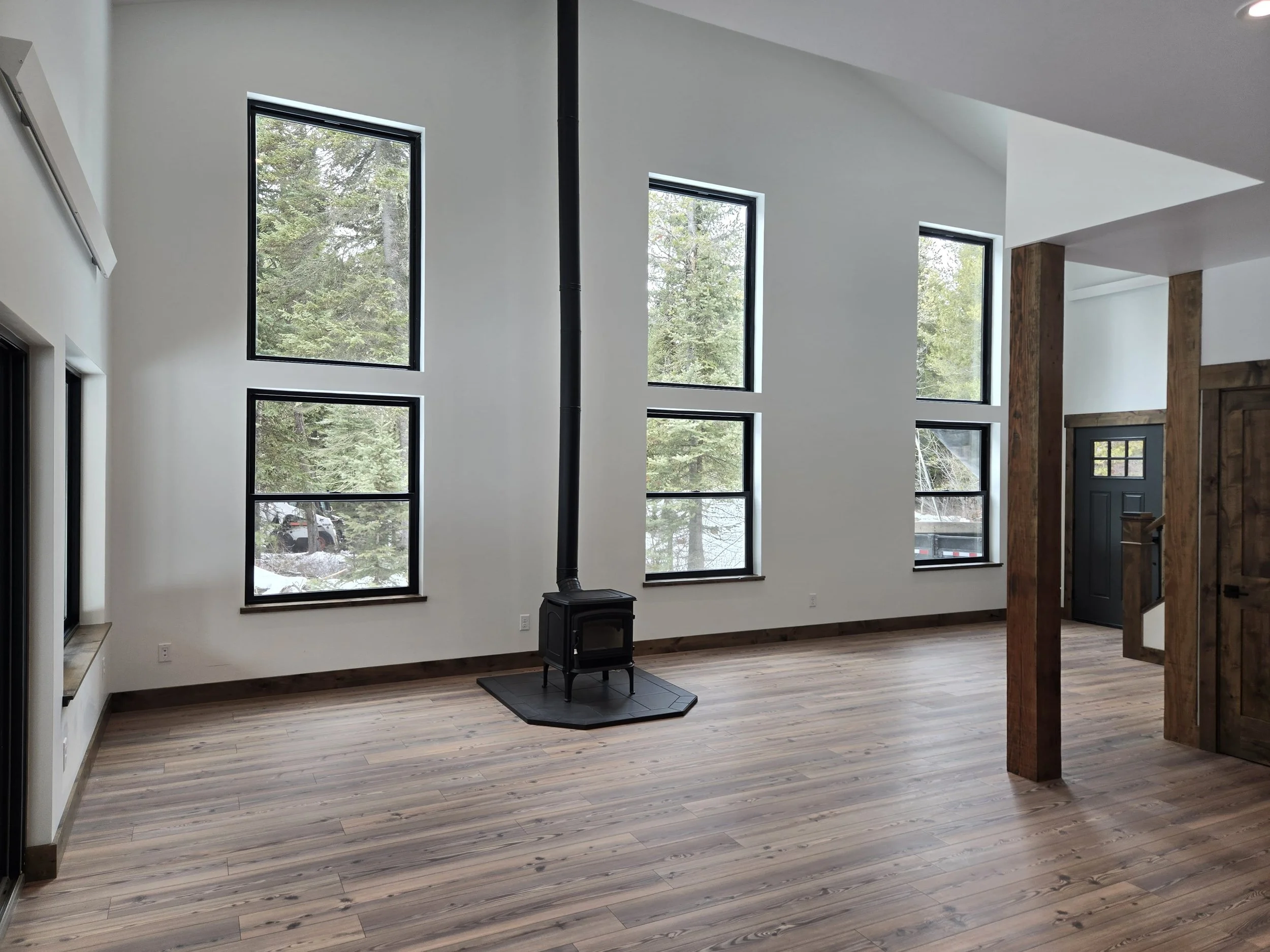 Empty living room with wooden flooring, white walls, multiple large windows, a wood stove, and a front door with a window.