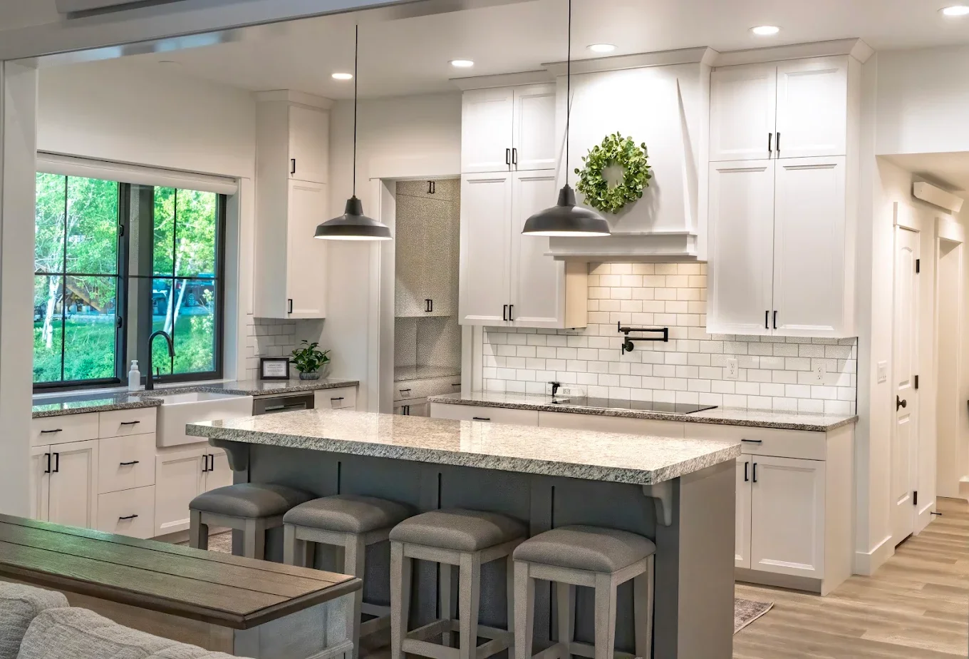 Modern kitchen with white cabinets, a large window over the sink, a granite island with four barstools, pendant lights, a tiled backsplash, and a decorative green wreath on the range hood in house in Star Valley Ranch, WY.