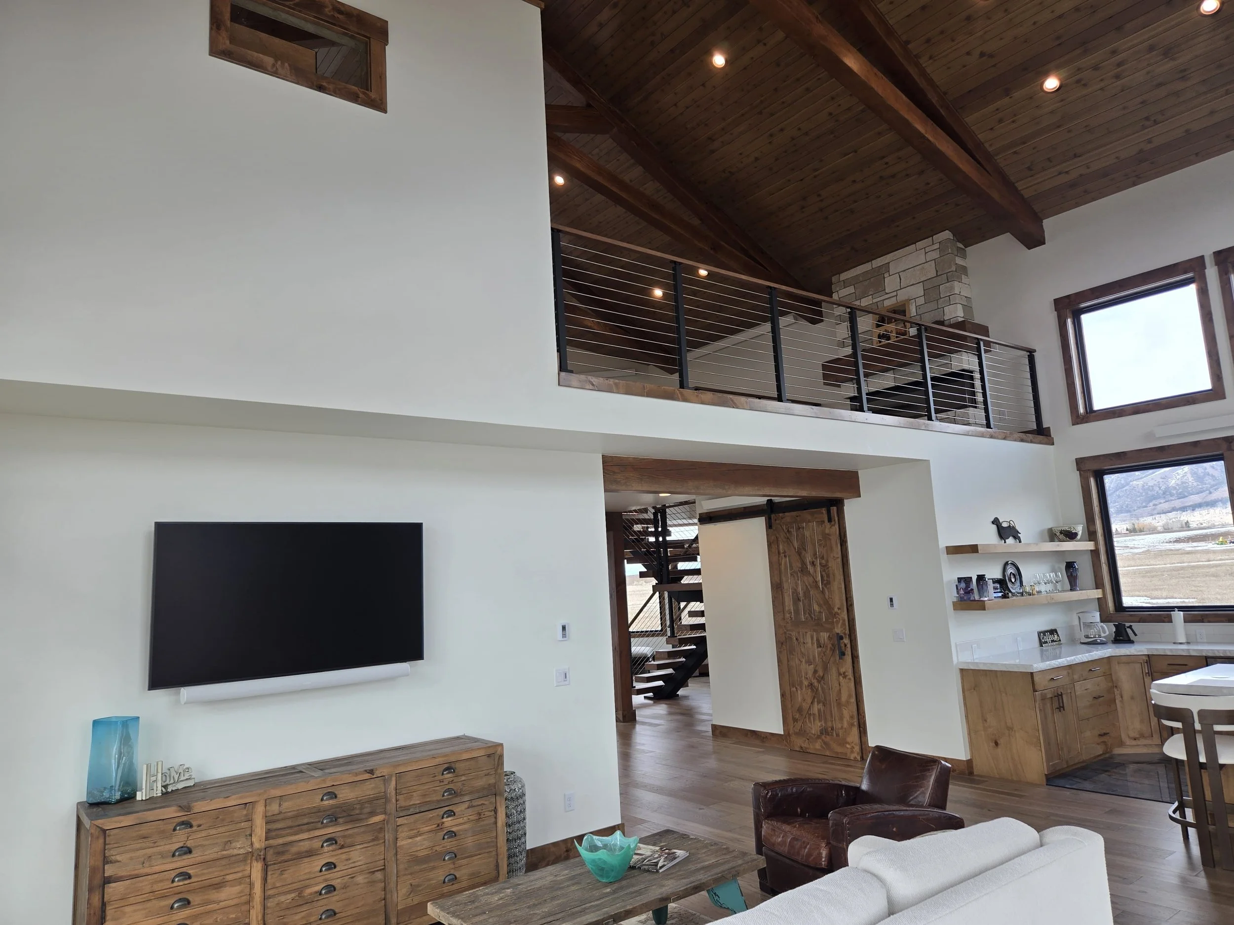 Living room with white walls, wooden ceiling and beams, large windows, a television on a wooden cabinet, and a loft with railing, stairs, and a fireplace.