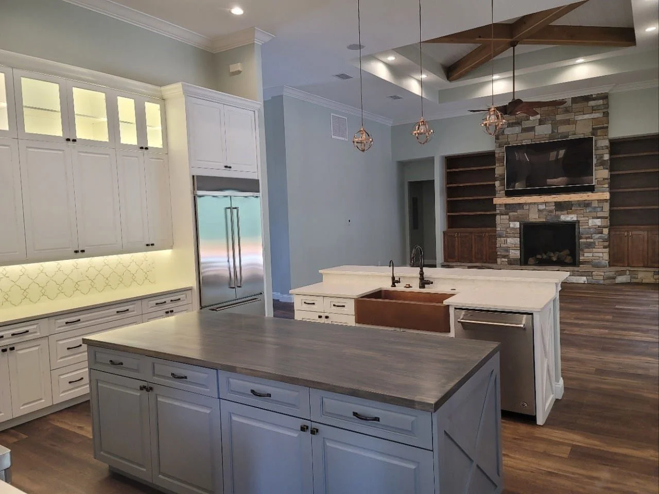 Modern open-concept kitchen with white cabinetry, a large gray island with a built-in sink, stainless steel refrigerator, and pendant lighting. In the background, there's a living area with a stone fireplace and a mounted TV, with wood flooring throu