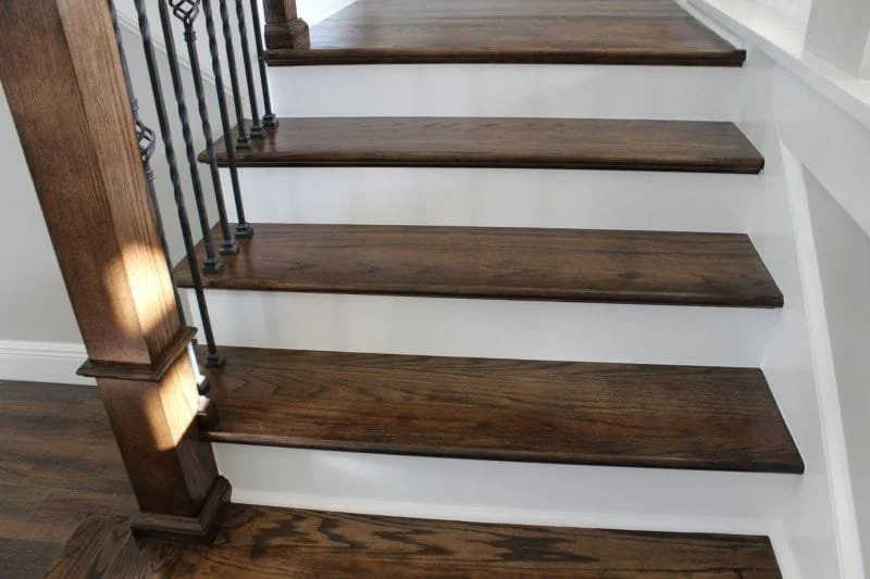 Close-up of a wooden staircase with dark brown steps and white risers, with a black metal railing and a wooden newel post on the left.