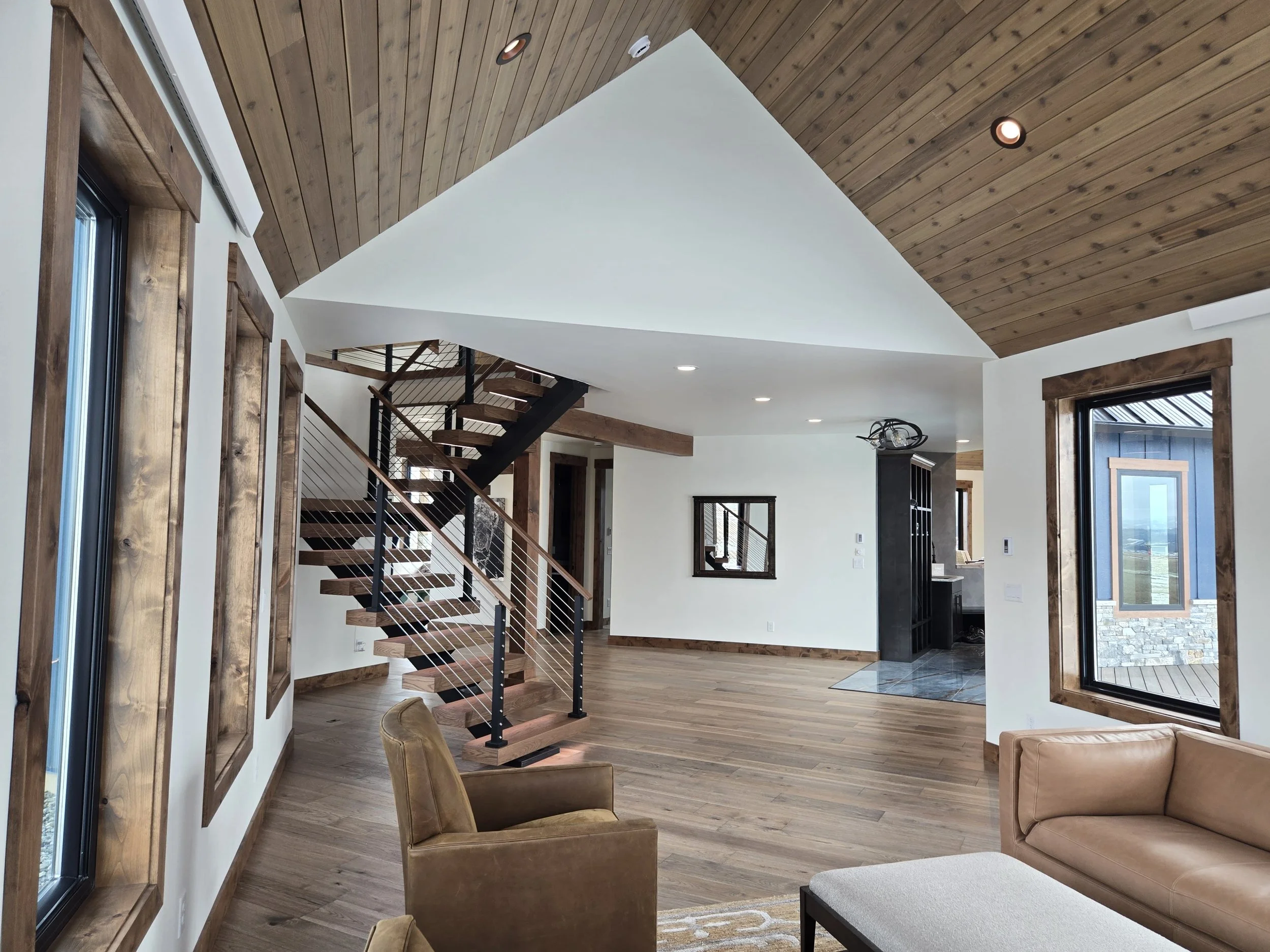 Interior of a modern home living room with wooden ceiling, large windows with wood trims, a staircase with wooden steps and black metal railing, and minimalistic furniture.
