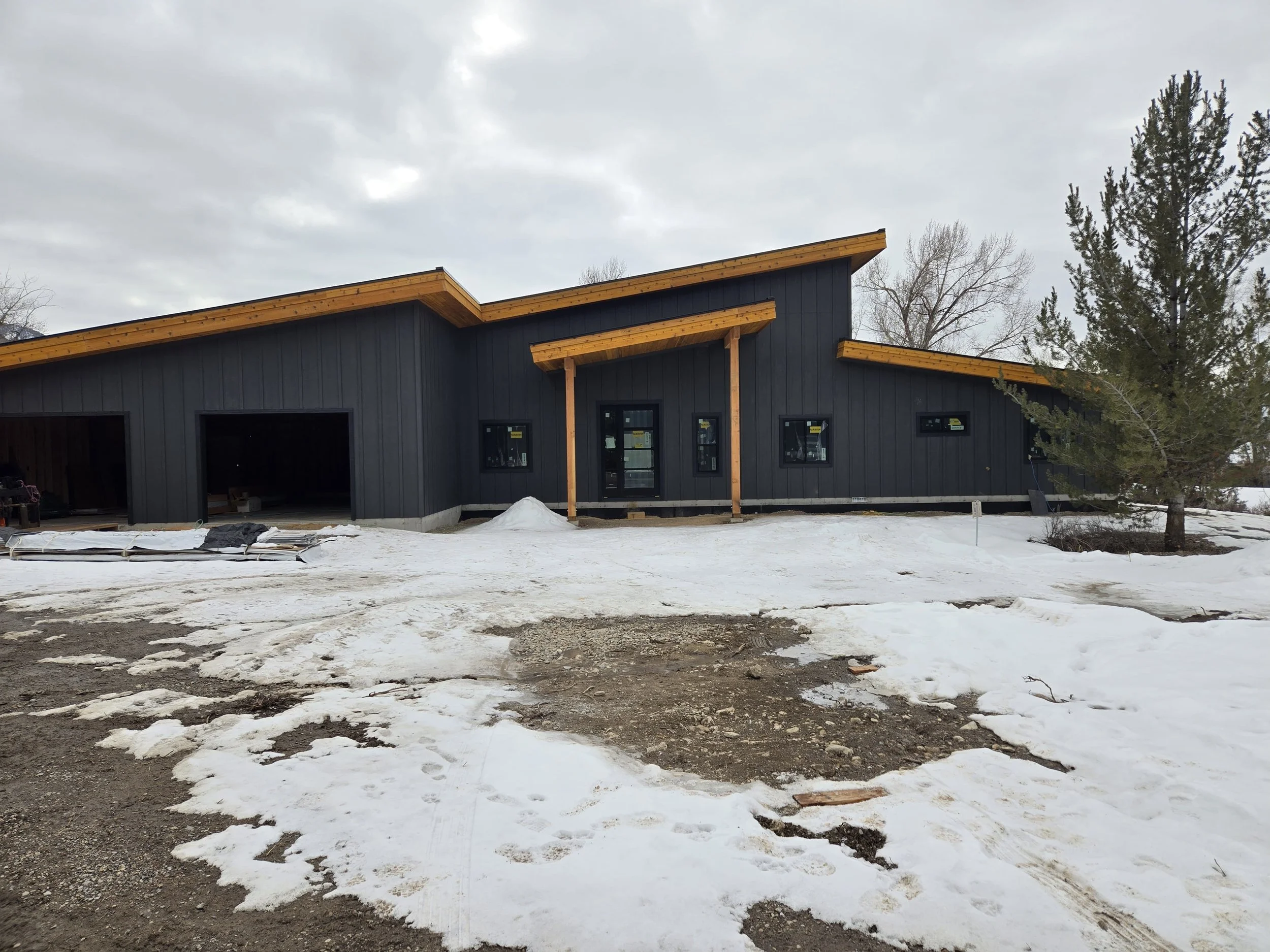 A modern house under construction with black exterior walls and unfinished driveway in a snowy landscape.
