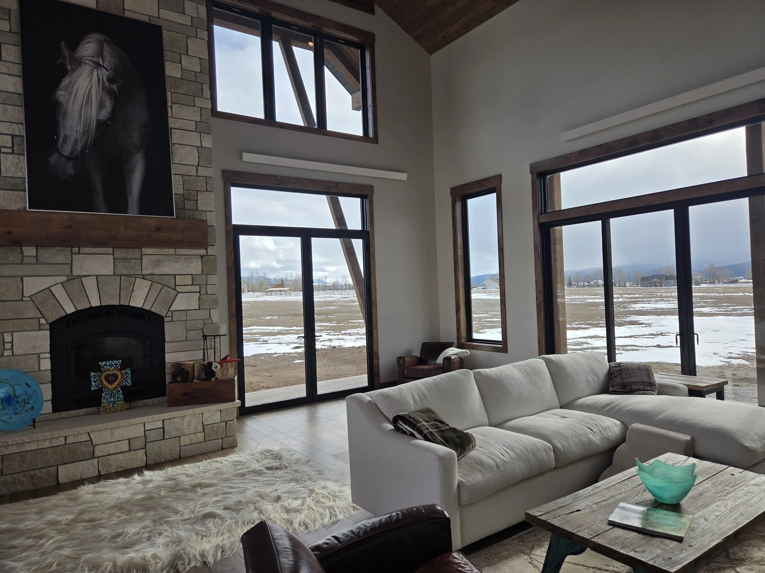 Living room with a white sectional sofa, a stone fireplace with a horse portrait above, large windows showing a snowy landscape outside, and a wooden coffee table.