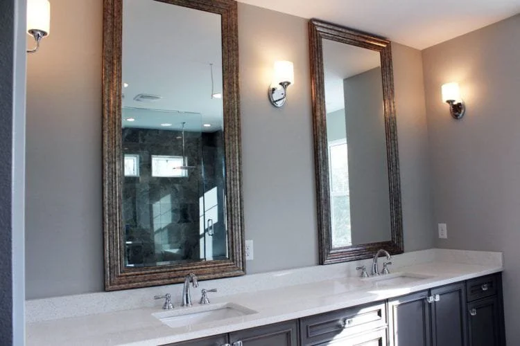 Bathroom vanity with two sinks, two large framed mirrors, three wall-mounted light fixtures, gray walls, and dark cabinetry.