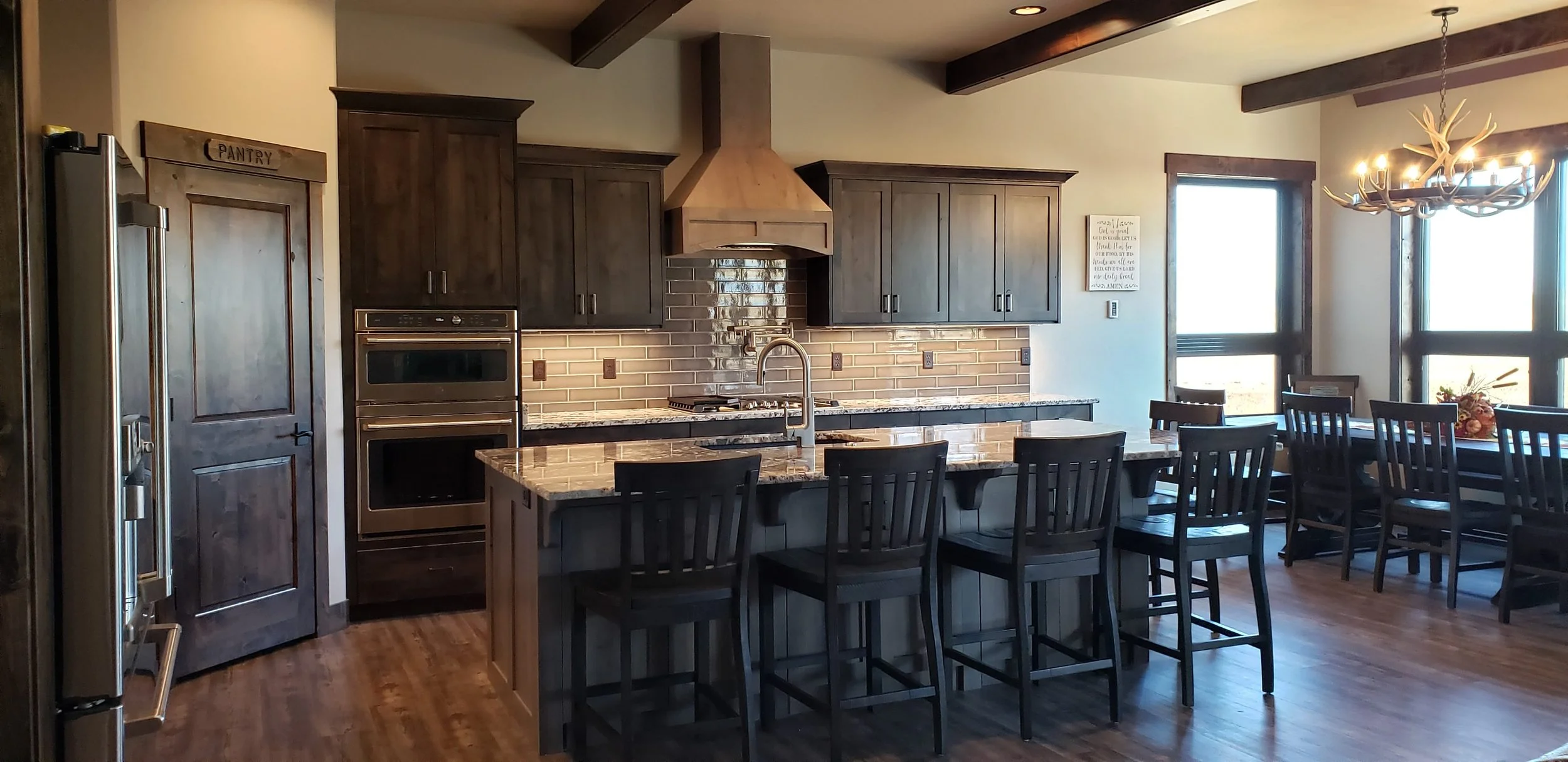 A spacious kitchen with dark wood cabinets, a granite island with four black chairs, a brick backsplash, a stainless steel refrigerator, double wall ovens, and a large dining table with chairs near windows with sunlight.