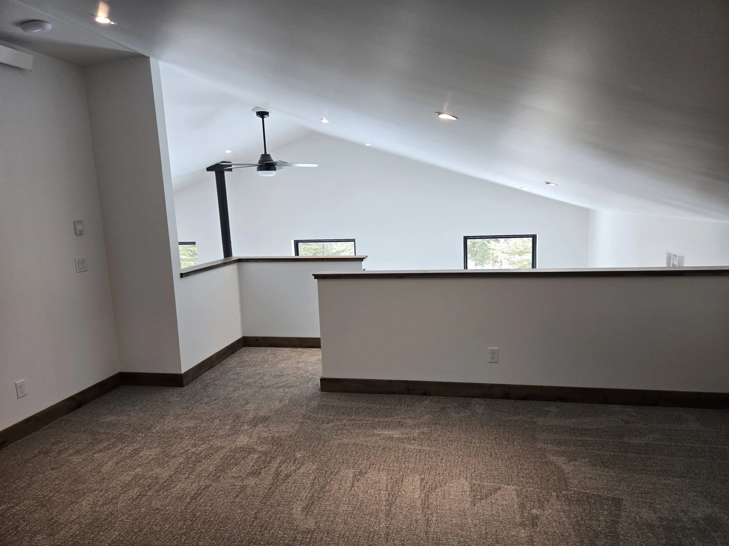Empty loft space with beige carpet, white walls, a black ceiling fan, and three windows letting in natural light.