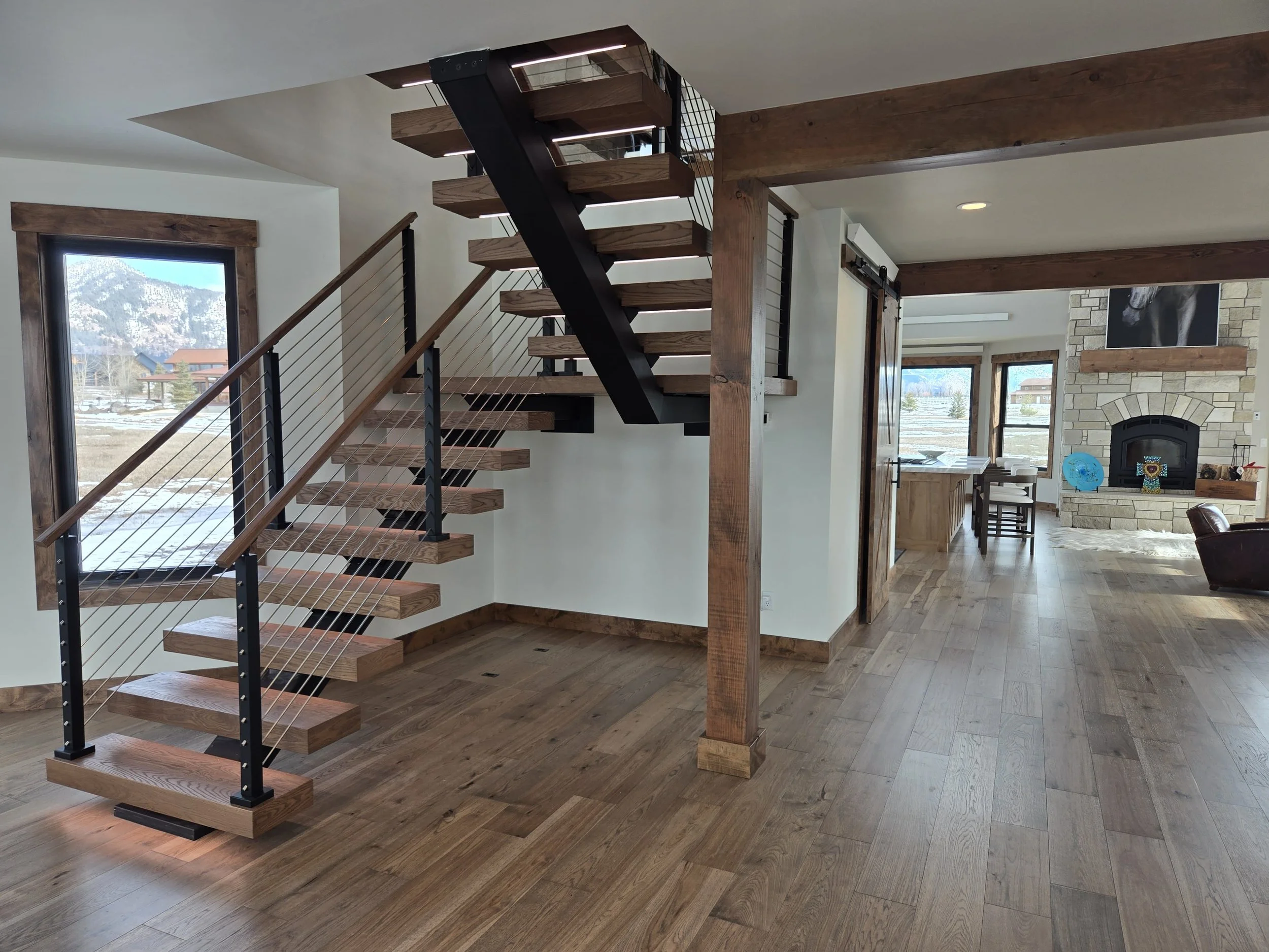 Interior of a modern home with wooden floors, staircase with wooden steps and metal railing, stone fireplace, and large windows with mountain scenery outside.