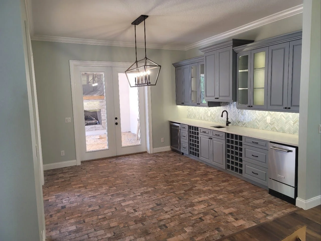 Empty kitchen with gray cabinets, brick-style flooring, and a glass door leading outside. A modern chandelier hangs from the ceiling.