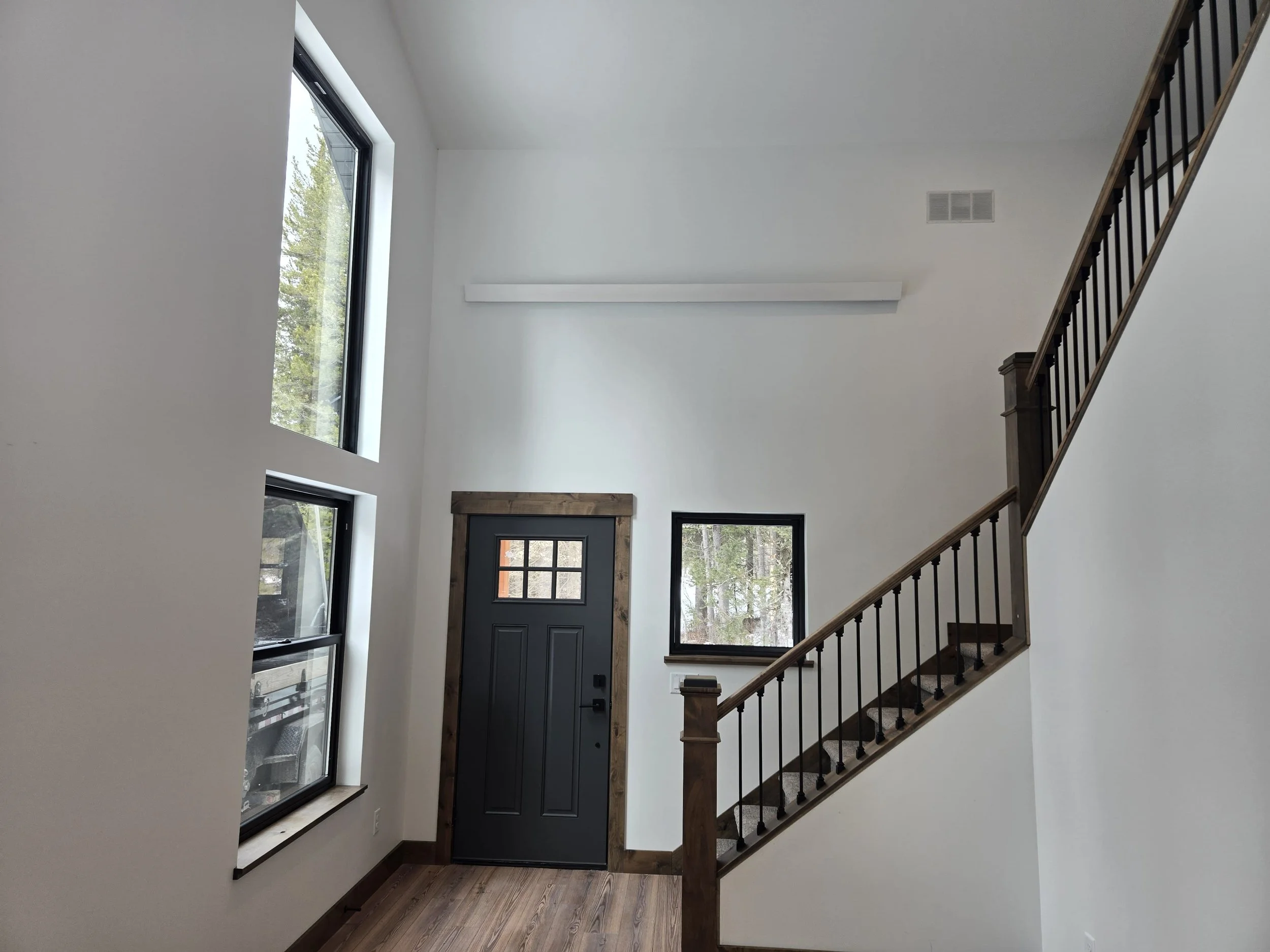 Interior view of a modern entryway with white walls, a black front door with a window, two large windows, a staircase with wooden railing and black balusters, and a wood floor.