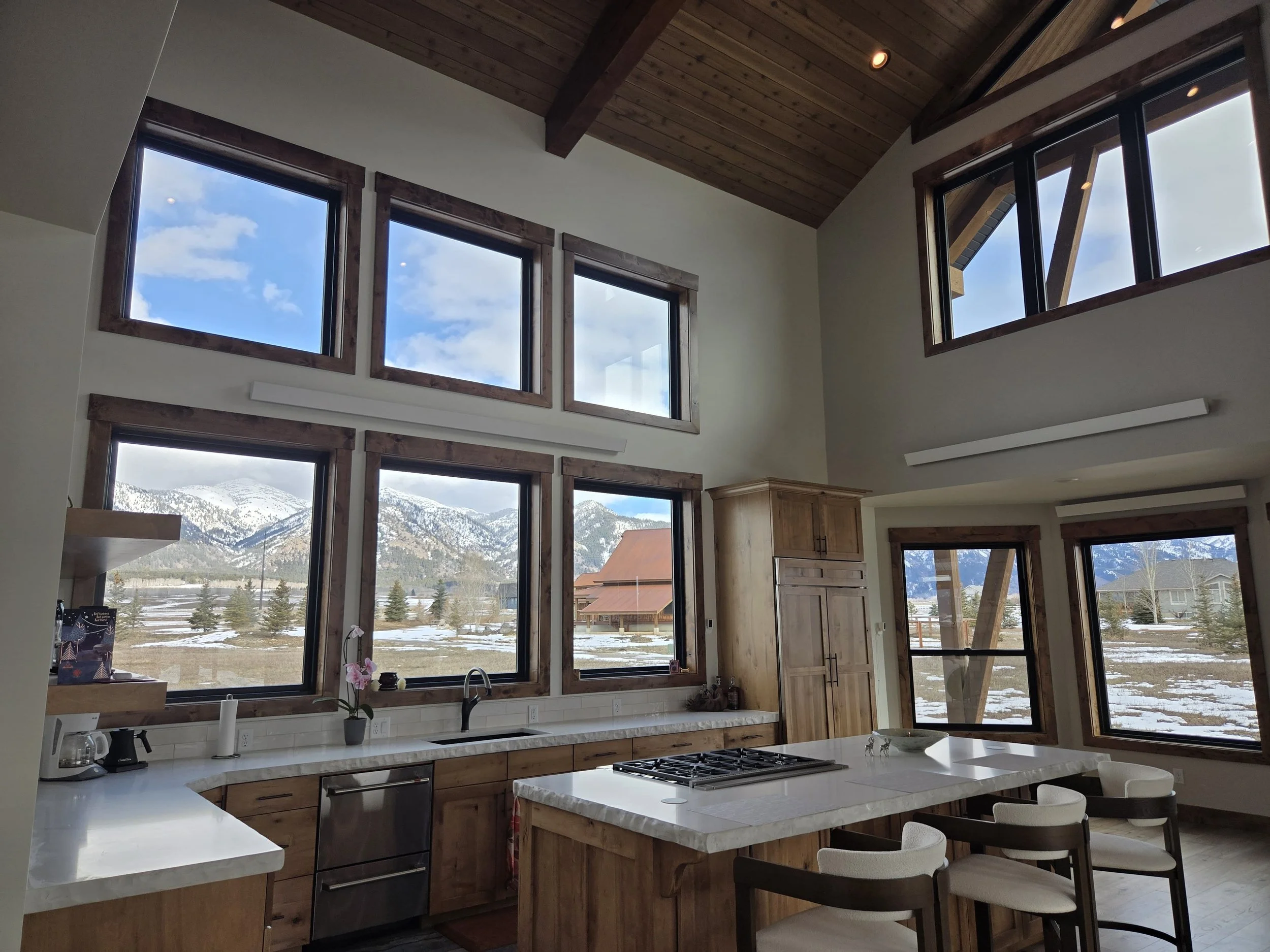 Bright kitchen with large windows showing snowy mountains and landscape outside, wooden cabinets, marble countertops, kitchen island with gas stove, chairs, and a sink.