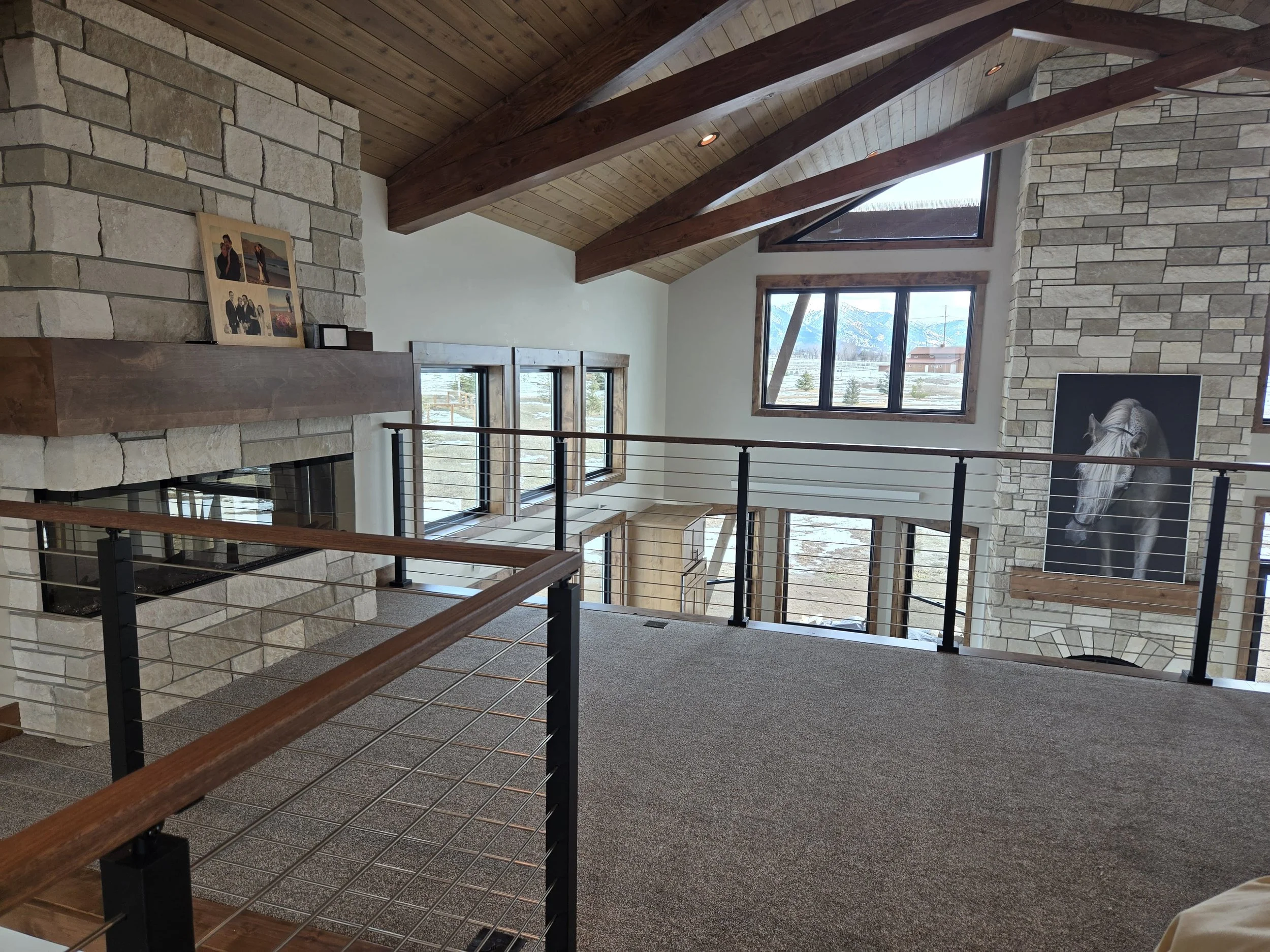 Interior of a modern home with high vaulted wooden ceilings, stone fireplace, large windows showing mountain views, and a black and white horse portrait on the wall.