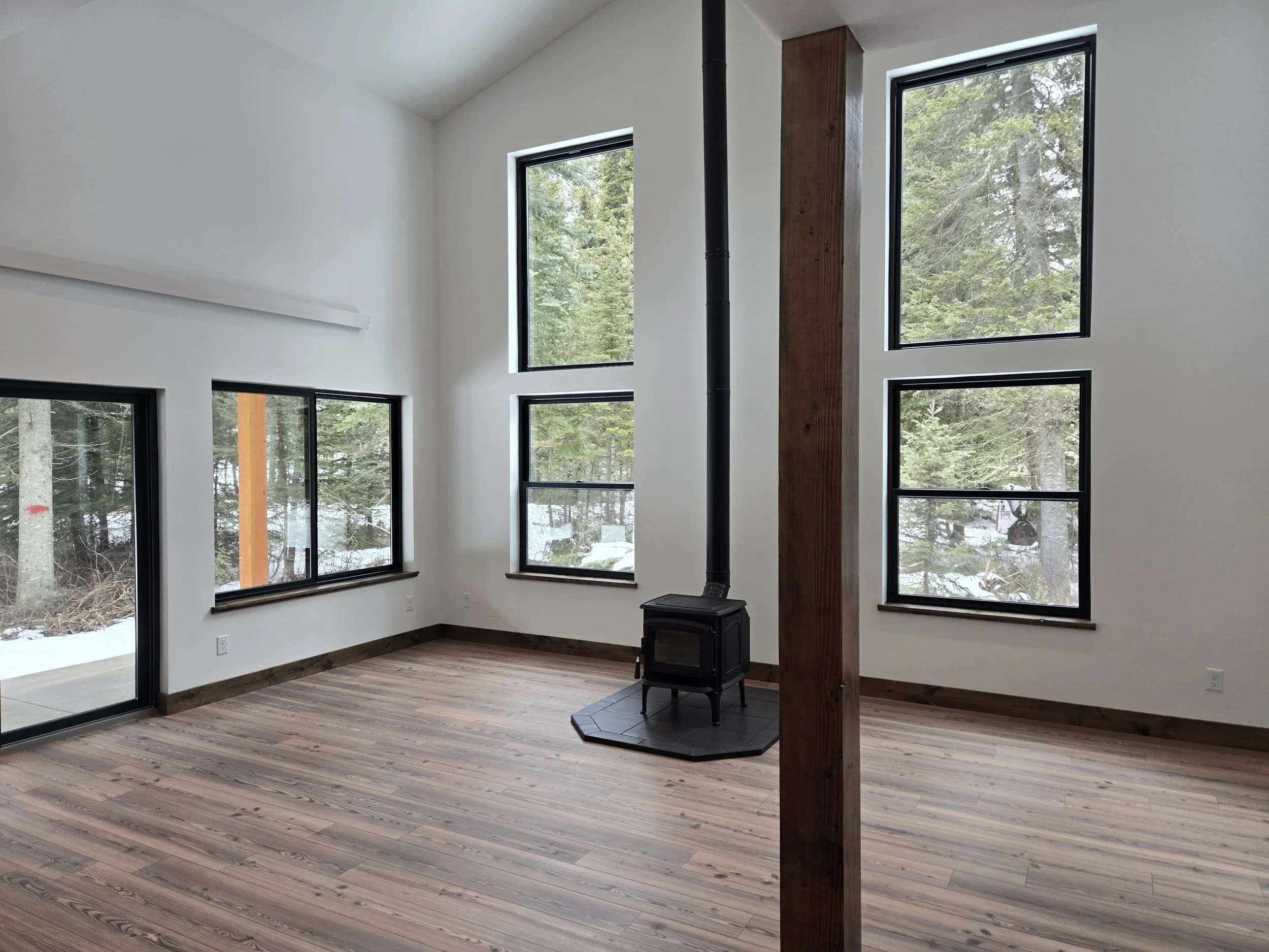 An empty room with large windows, a wood stove, and wooden flooring, overlooking trees outside.
