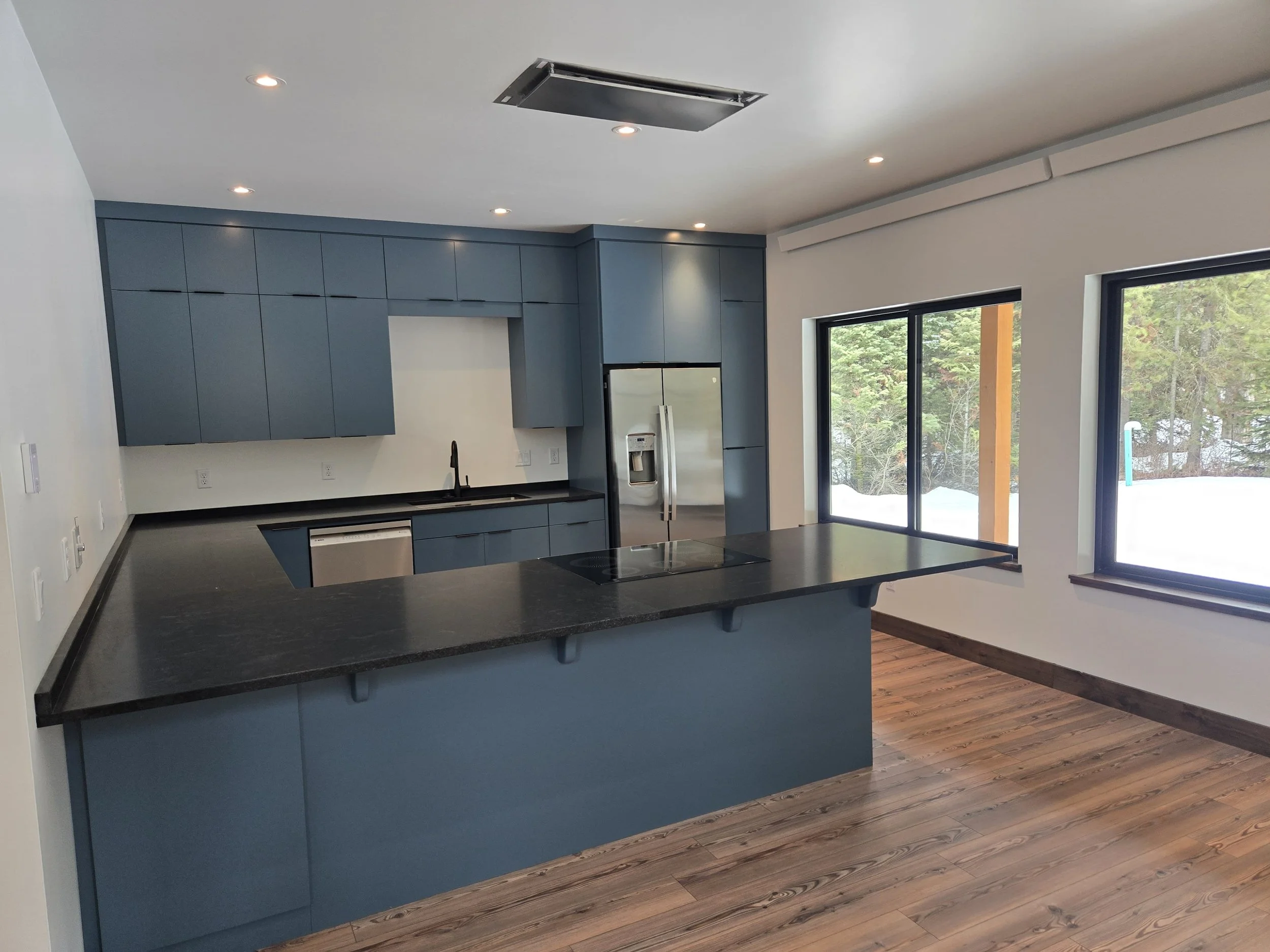 Modern kitchen with blue cabinets, black countertops, stainless steel refrigerator, and large windows overlooking a snowy landscape.