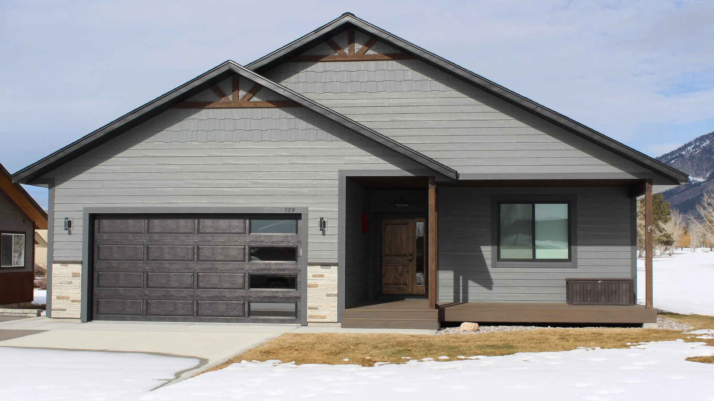 Modern gray house with dark garage door and front porch, snow on ground, mountain in background.