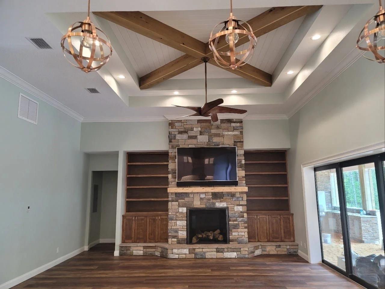 Living room with a stone fireplace, wooden built-in shelves, a mounted flat-screen TV, wood ceiling beams, modern pendant light fixtures, a ceiling fan, hardwood flooring, and a sliding glass door leading outside.
