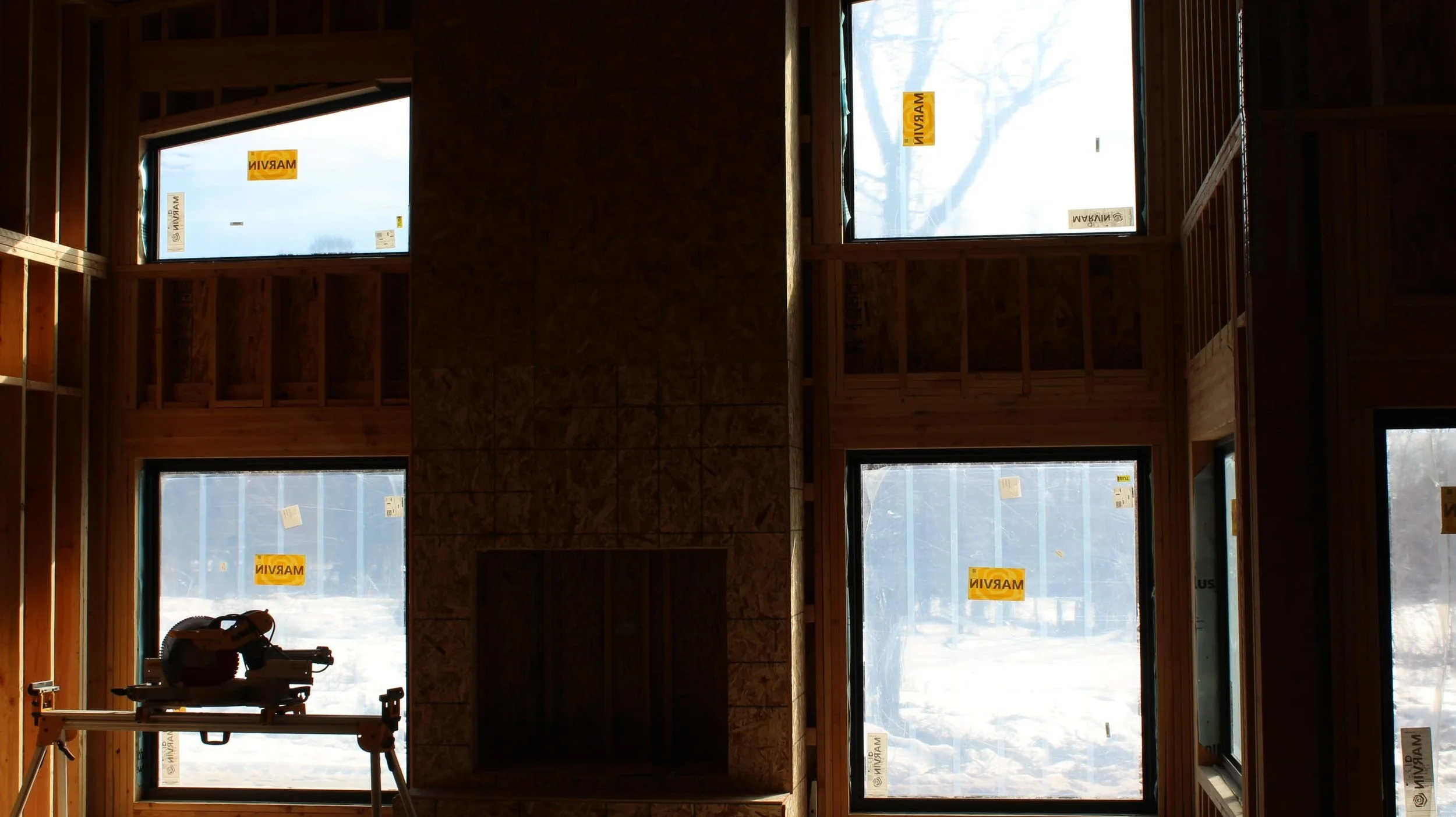 Interior of a framed building under construction, showing wooden wall studs and large window openings with snowy outdoor landscape visible, labeled 'MARVIN' on the window stickers.