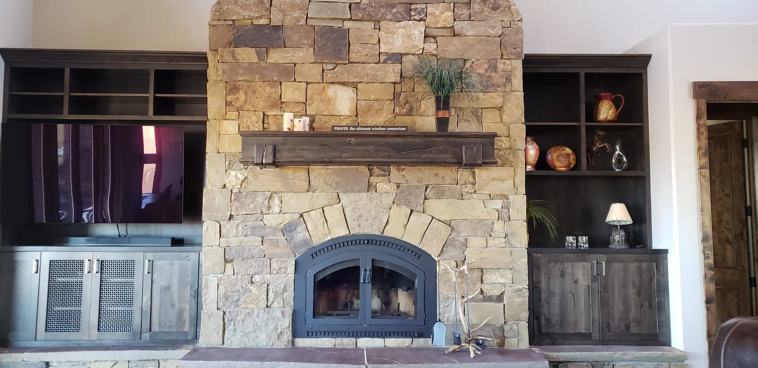 Living room with a stone fireplace, black metal firebox, and dark wood built-in shelves on either side with decorative vases and a lamp.