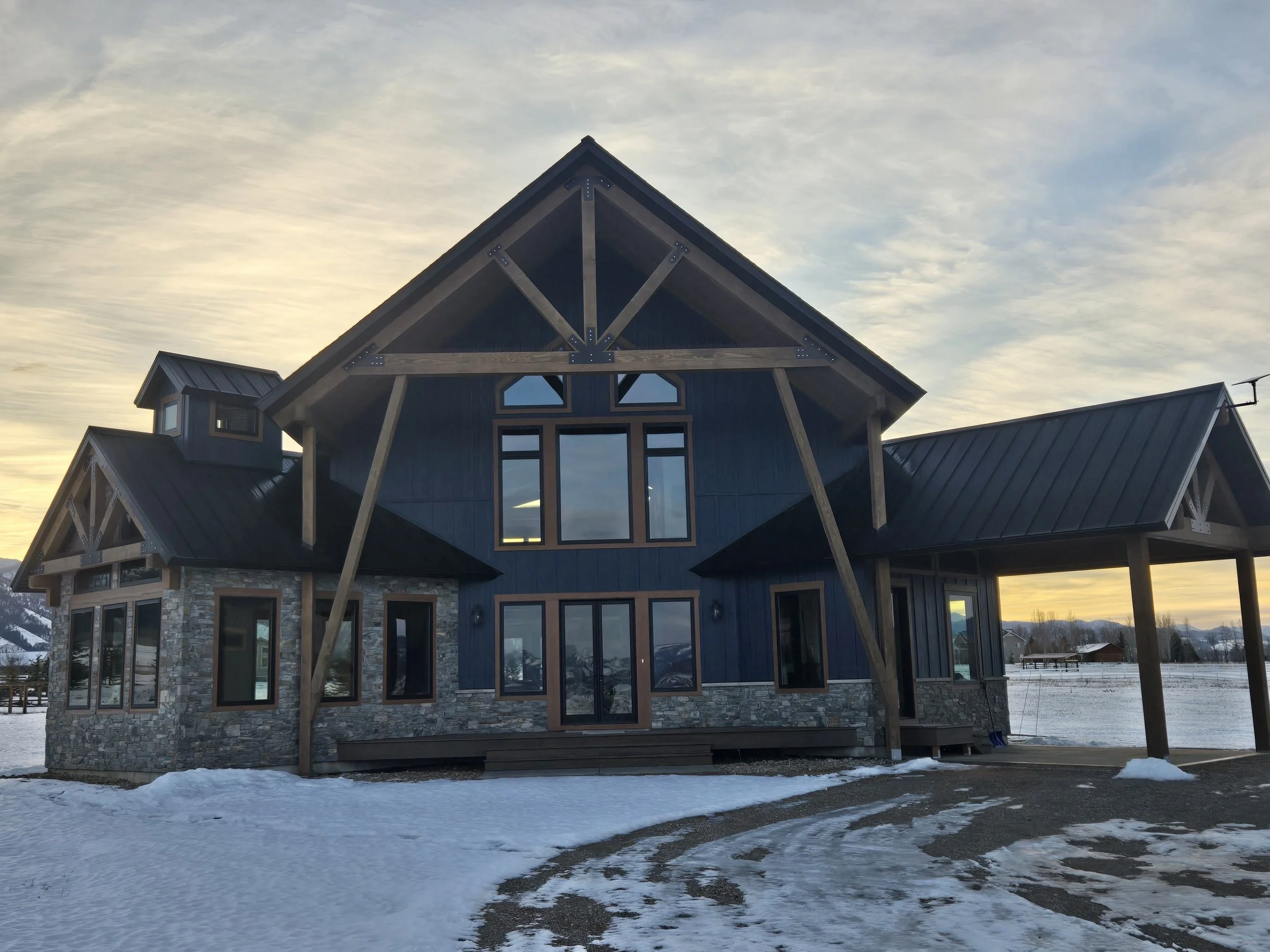 A modern house with a stone lower exterior, dark blue upper siding, and metal roof, set against a snowy landscape at dusk.