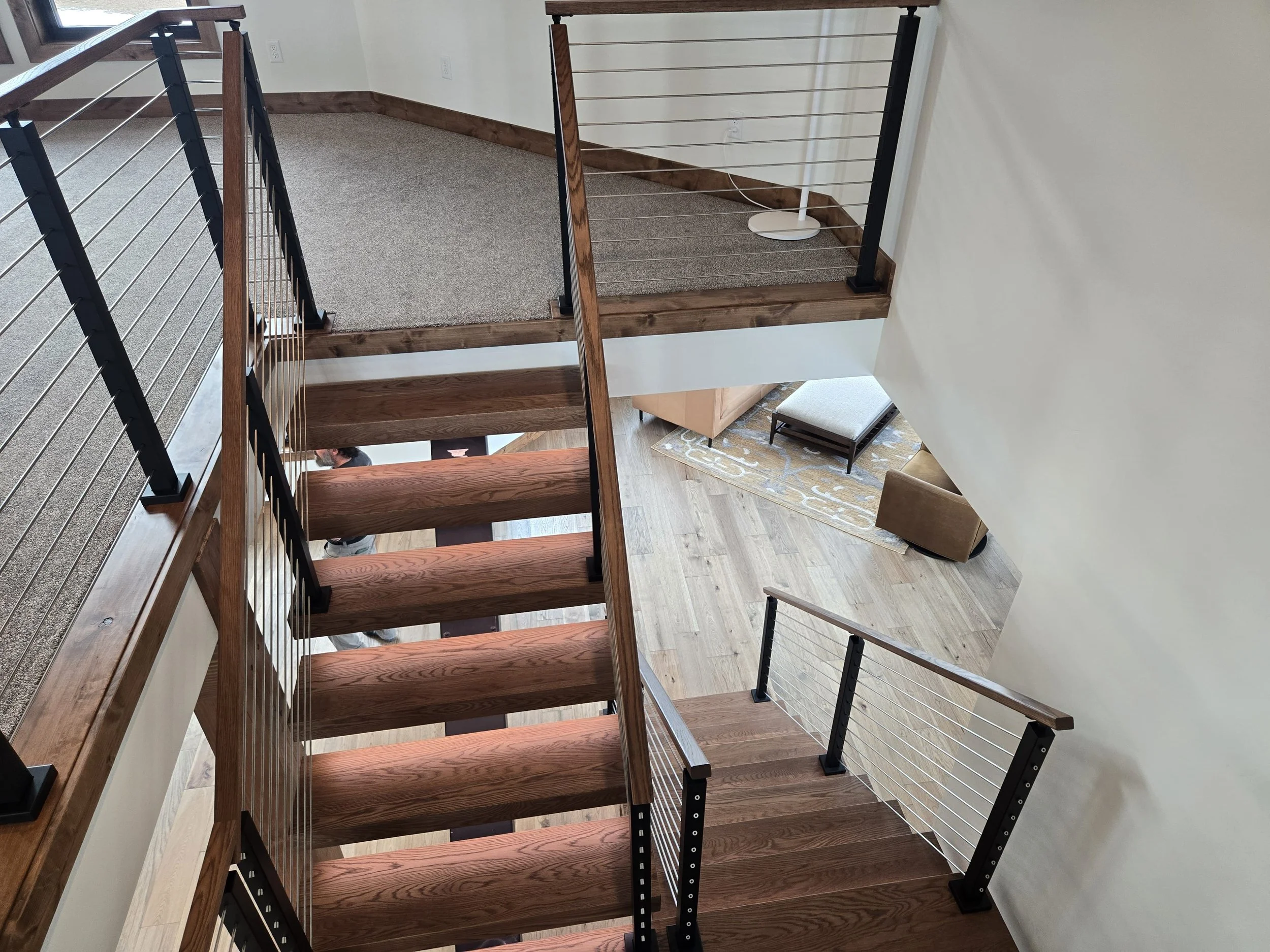 View of a staircase with wooden steps and metal railings, leading to a lower living area with a beige armchair, a coffee table, an area rug, and a wooden floor.