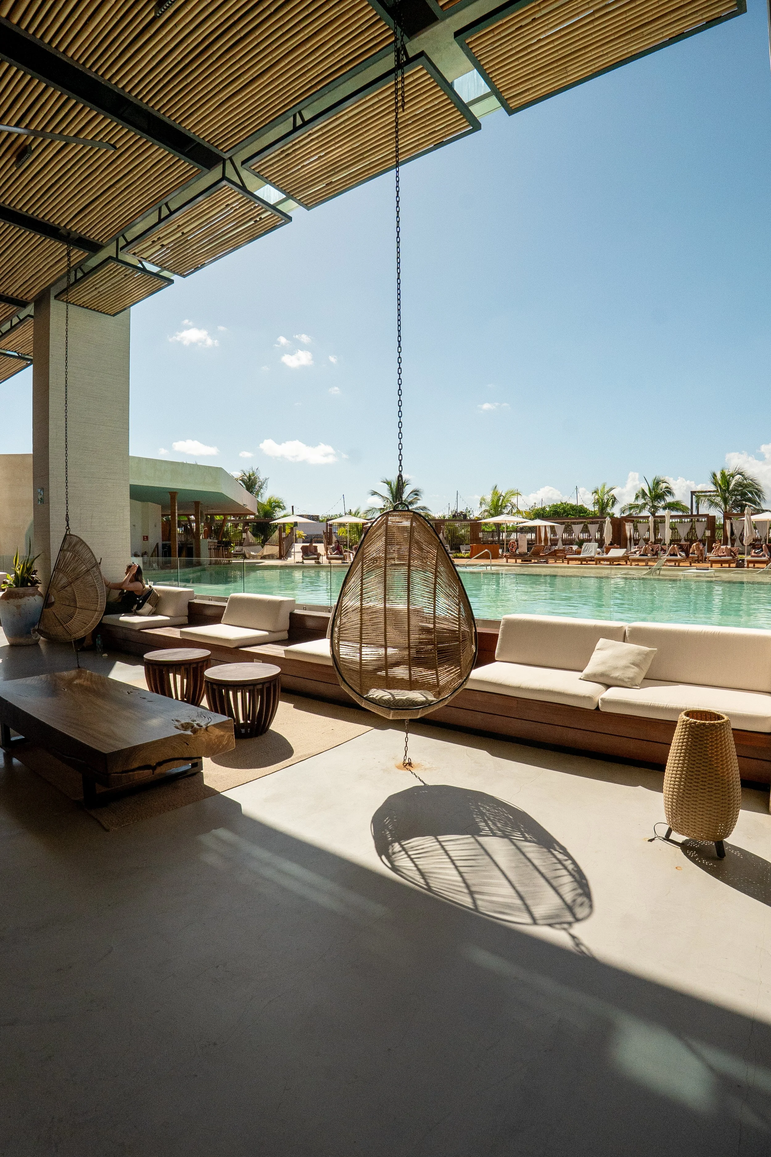 A poolside lounge area with hanging wicker chairs, sofas, and tables, overlooking a swimming pool under a clear blue sky, with palm trees in the background.