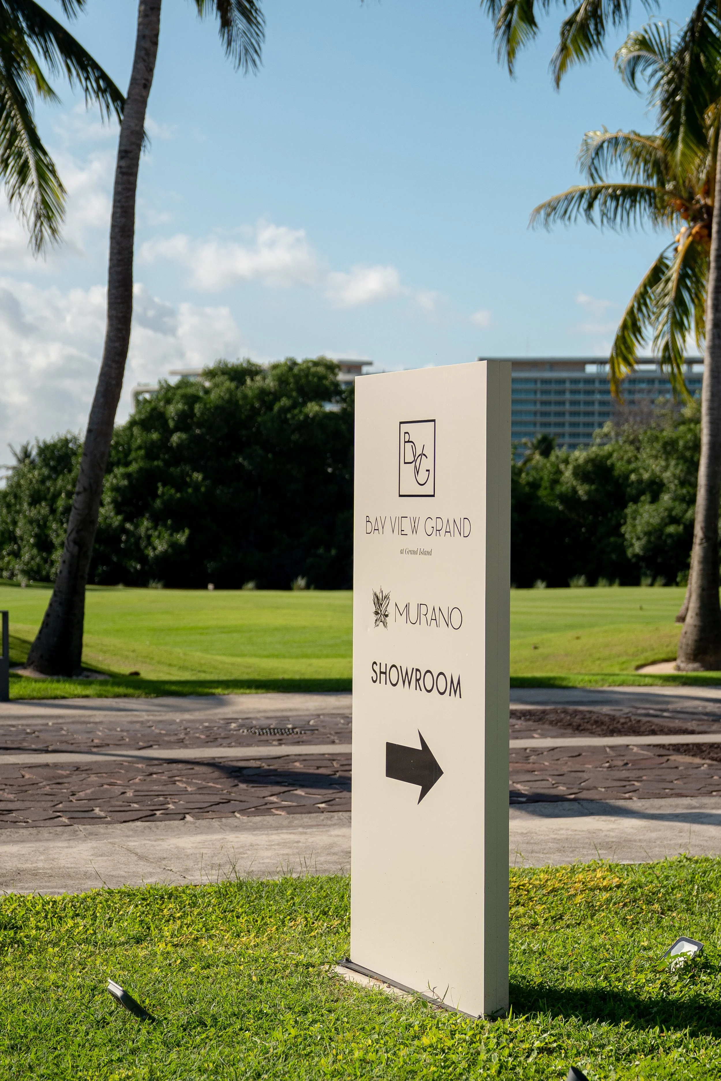 A white sign with black text and an arrow pointing to the right, located outdoors in a park-like setting with palm trees, grass, and a building in the background. The sign reads "Bay View Grand at Ground Island," "Murano," and "Showroom."