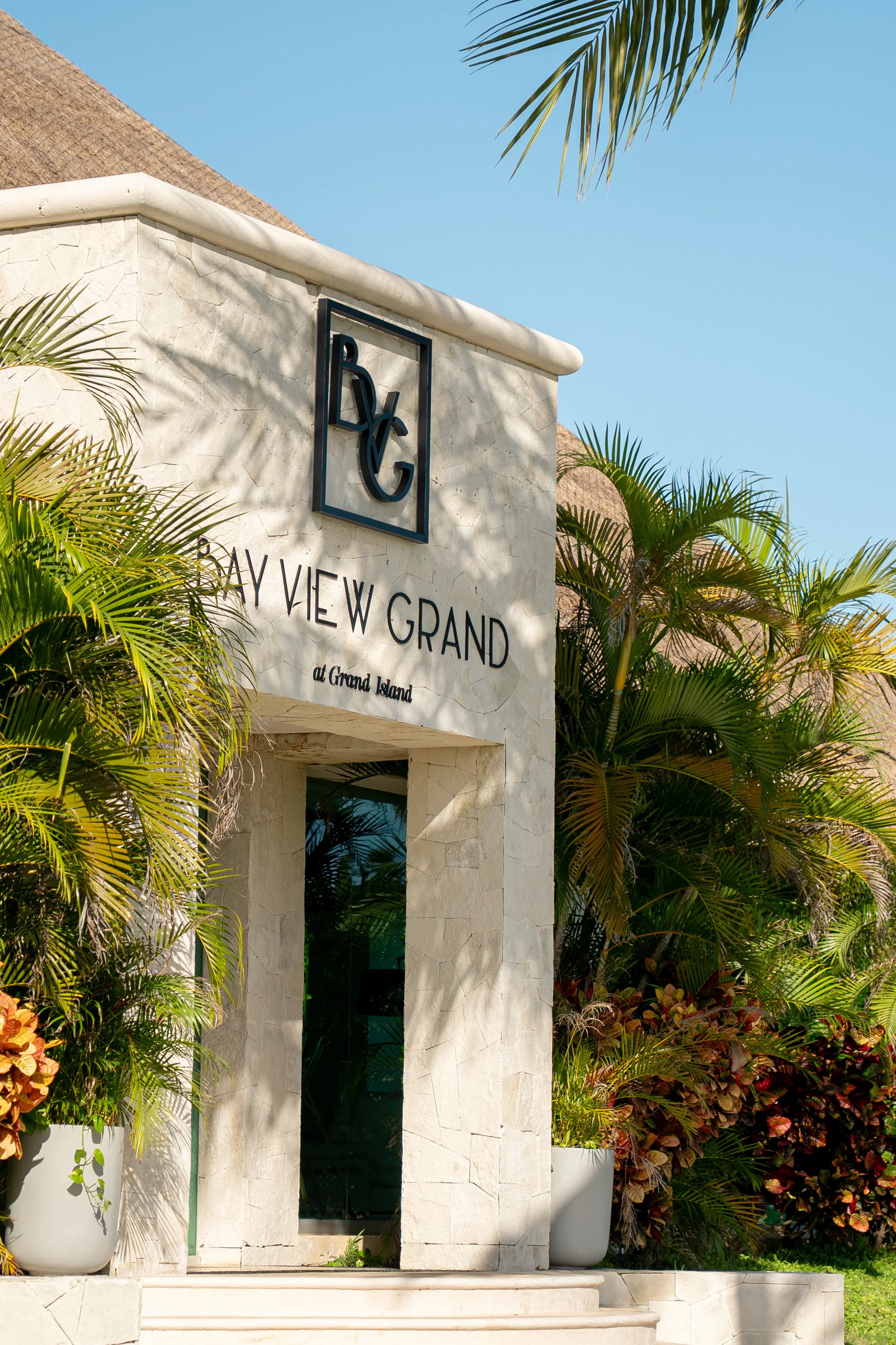 Entrance sign for Bay View Grand at Grand Island with lush tropical plants and clear blue sky.