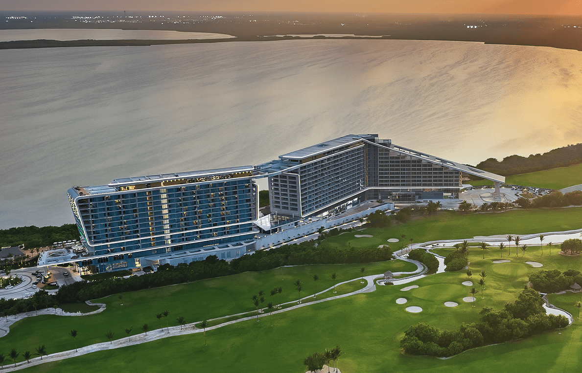 Aerial view of a modern hotel building near water and a golf course at sunset.