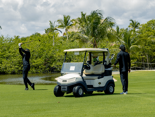 Two men on a golf course, with one swinging a golf club and the other standing near a golf cart, surrounded by palm trees and greenery.