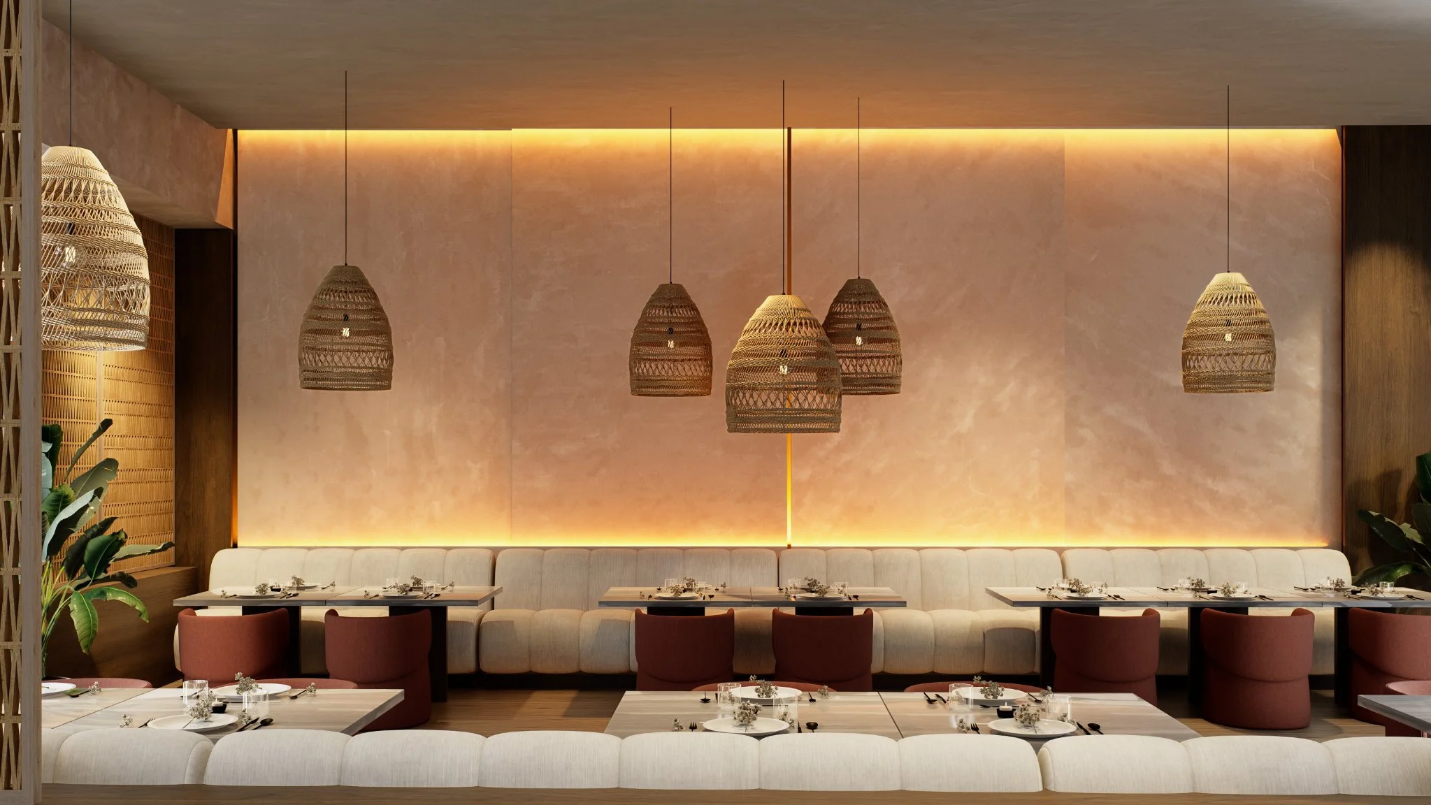 Interior of a restaurant with soft lighting, hanging wicker pendant lamps, a long beige upholstered banquette, round and rectangular tables set with glassware, plates, silverware, small floral centerpieces, and pink chairs. There are lush green plant