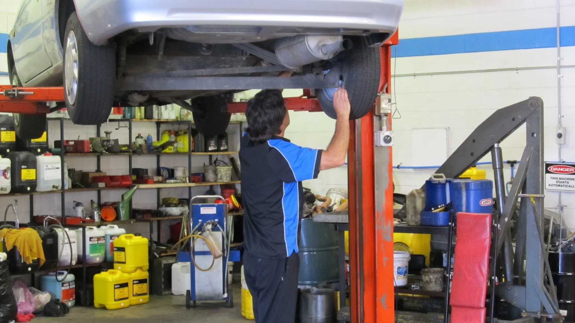Mechanic inspecting underside of a silver car lifted on a hydraulic lift in an auto repair shop.