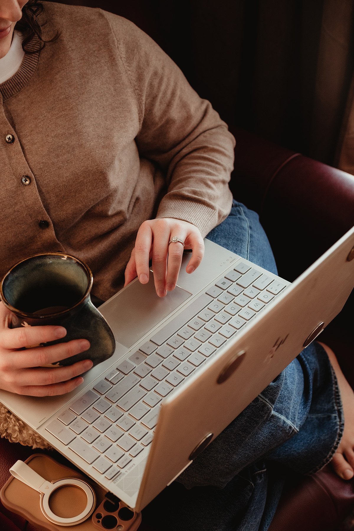 Person wearing a beige sweater working on a silver laptop, holding a black mug, sitting on a leather seat.