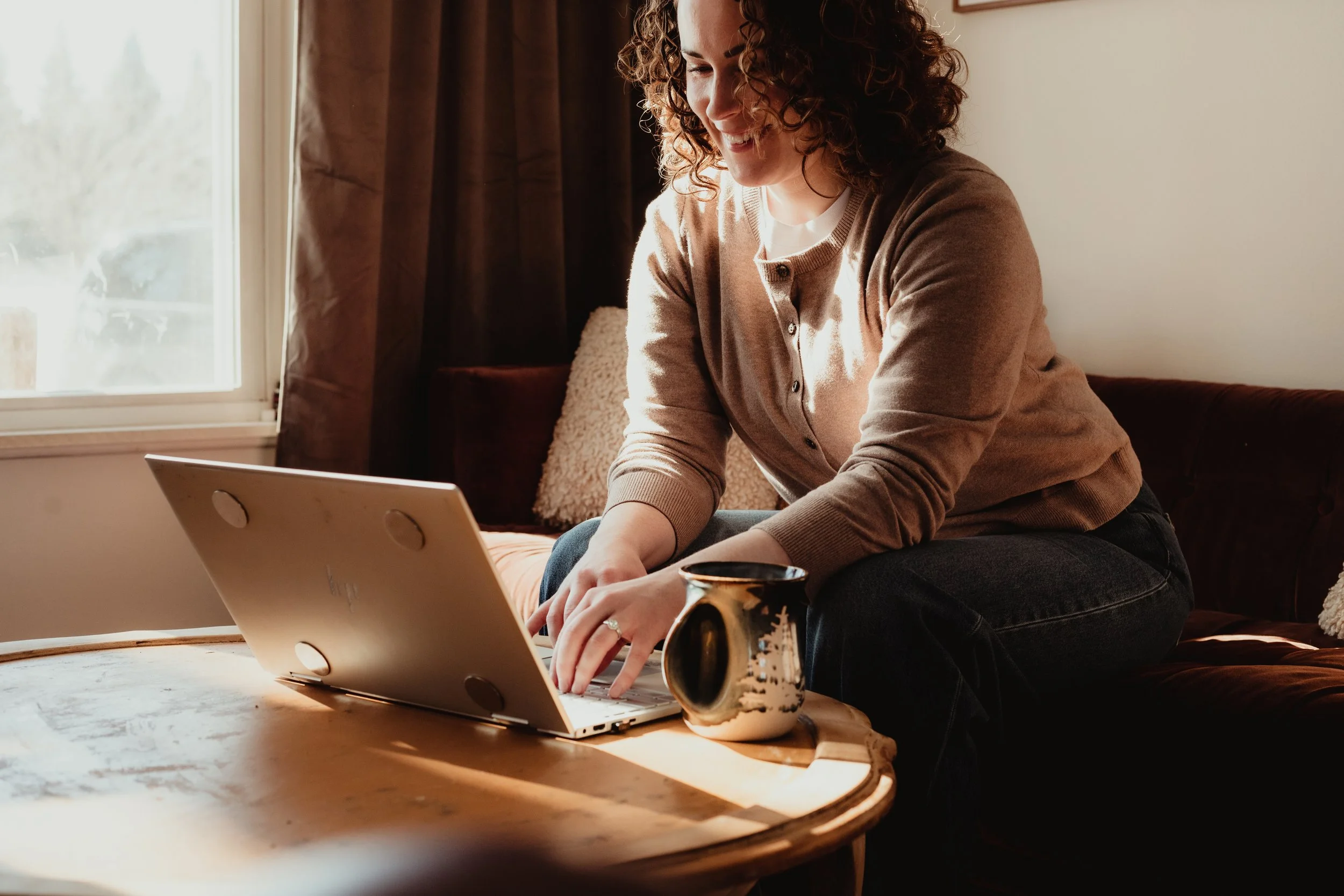 A woman with curly hair sitting on a couch, smiling and using a laptop on a wooden coffee table inside a room with a window and curtains.