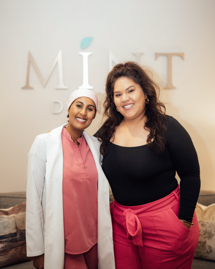 Two women standing together, smiling, in front of a wall with the word 'MINT' displayed. One woman is wearing medical scrubs and a white coat, the other is in a black top and pink pants.