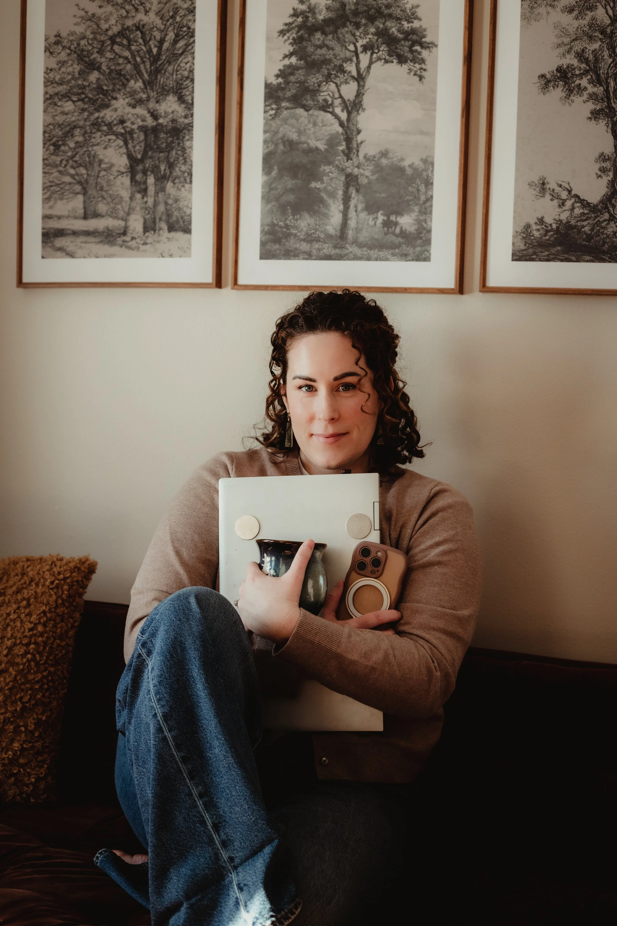 A woman with curly hair sitting on a couch, holding a laptop, a coffee mug, and a smartphone. Behind her are three framed black-and-white landscape pictures of trees on the wall.