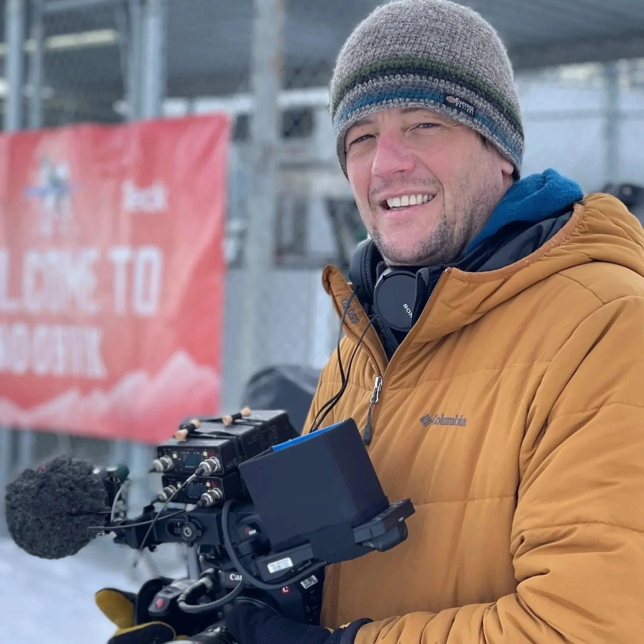 A man wearing a brown jacket, gray beanie, and headphones around his neck holding a camera with a furry microphone cover.