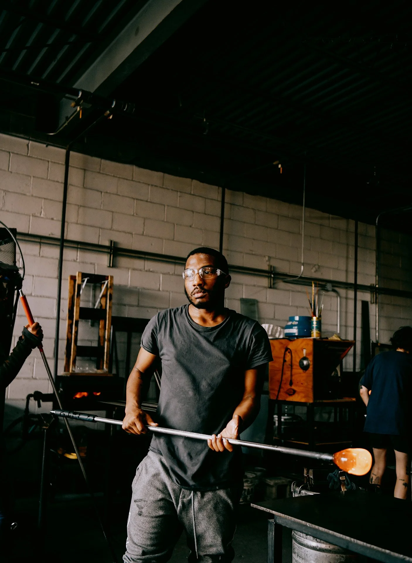 A man with glasses holding a long metal rod with a glowing orange tip, standing in an industrial-style room with exposed brick walls and black ceiling pipes.